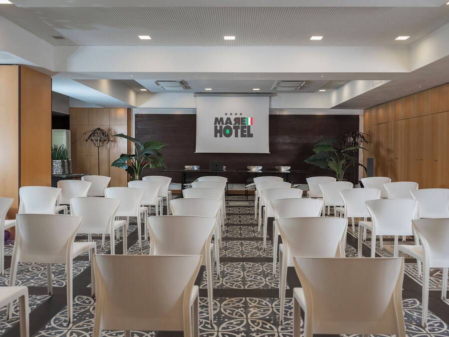 A conference room with rows of white chairs, a large screen displaying "MARE HOTEL" logo, and a patterned carpet.