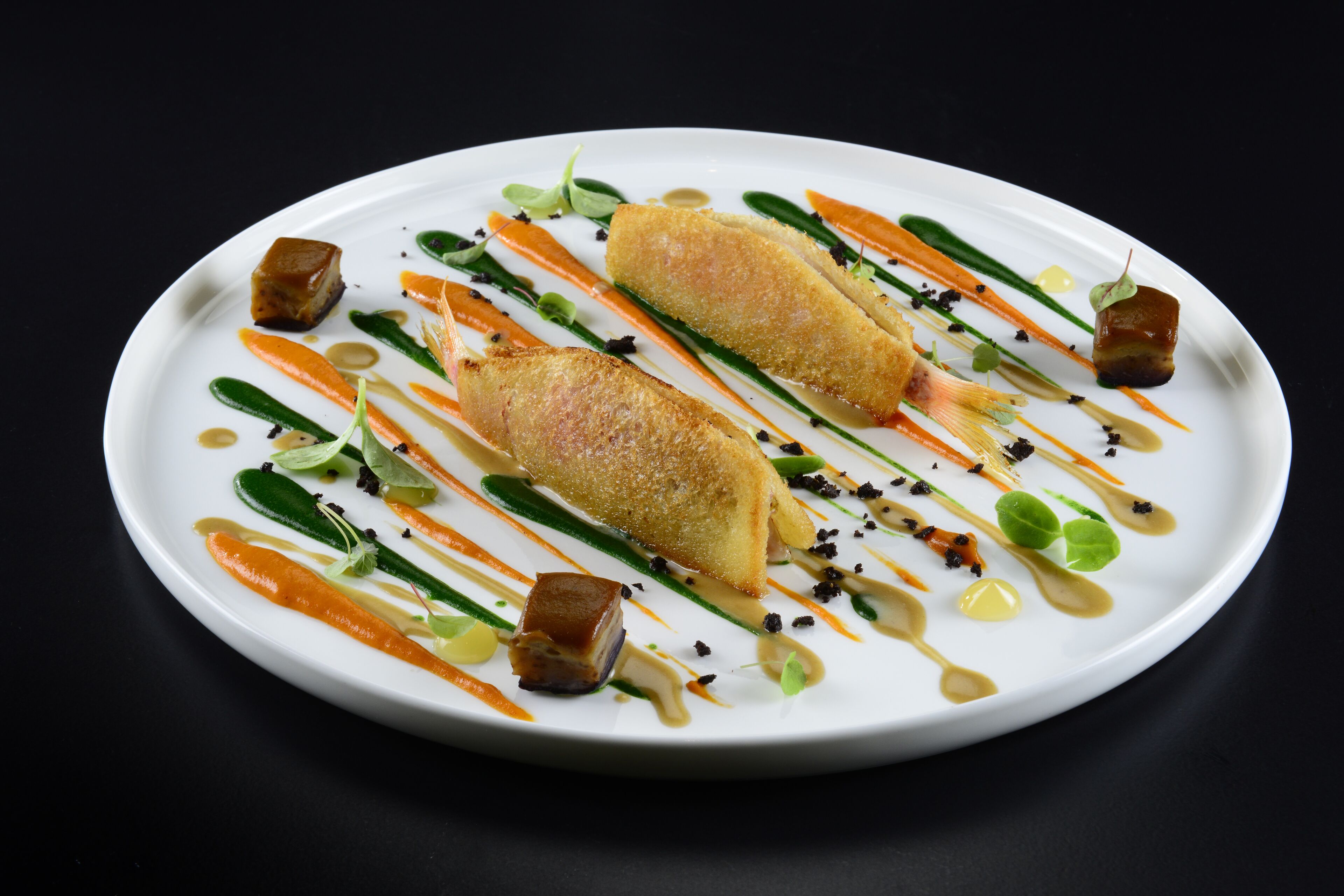 Un plato blanco con dos croquetas doradas, verduras verdes y pequeños cubos de un alimento oscuro, todo sobre un fondo negro.