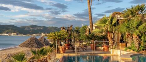 Un área de piscina con palmeras, vista a la playa y una cabaña de techo de paja.