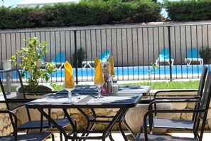 Un área de comedor al aire libre con vista a la piscina, con una mesa dispuesta con servilletas amarillas, saleros y pimenteros, y un vaso de jugo de naranja.