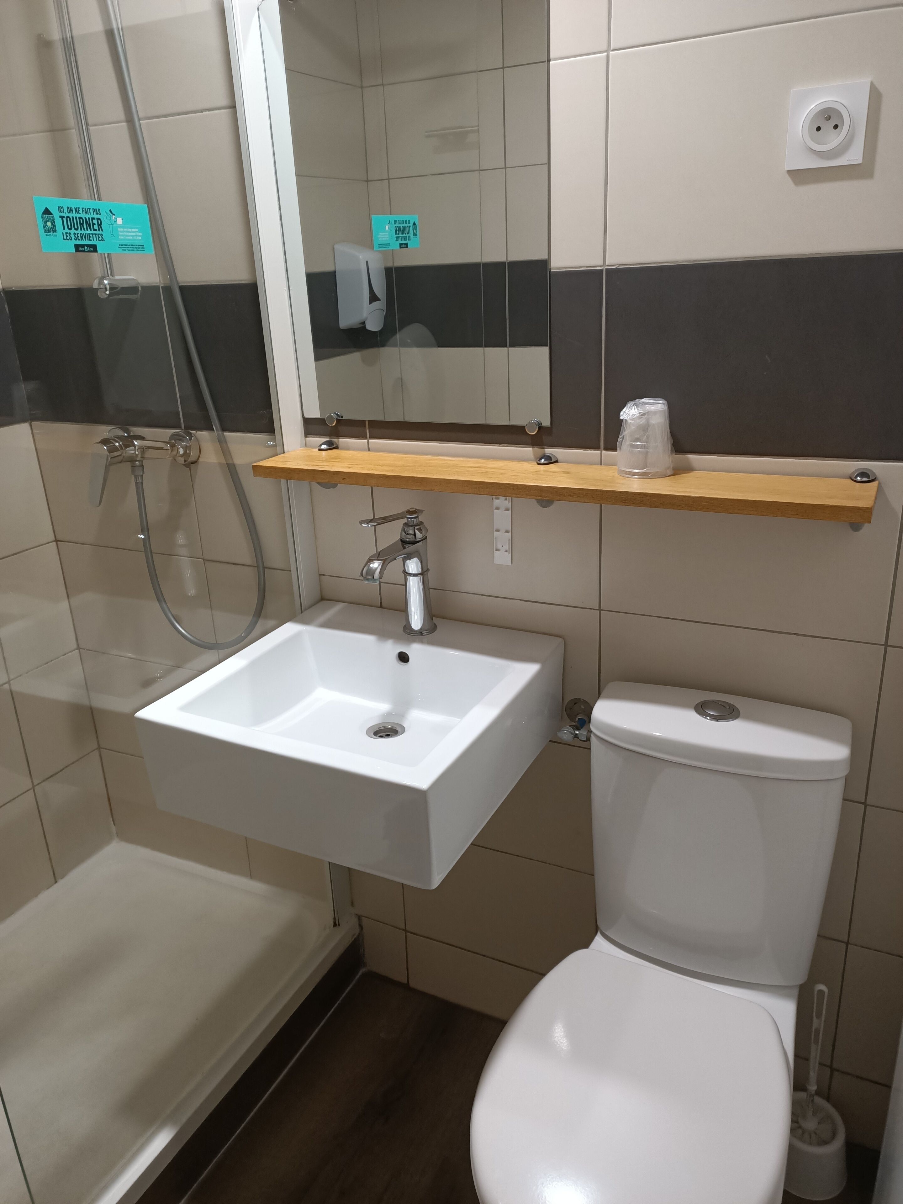 A bathroom with a white sink, a toilet, a shower, and a mirror. There is a sign on the wall and a shelf above the sink.