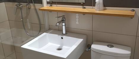 A bathroom with a white sink, a toilet, a shower, and a mirror. There is a sign on the wall and a shelf above the sink.