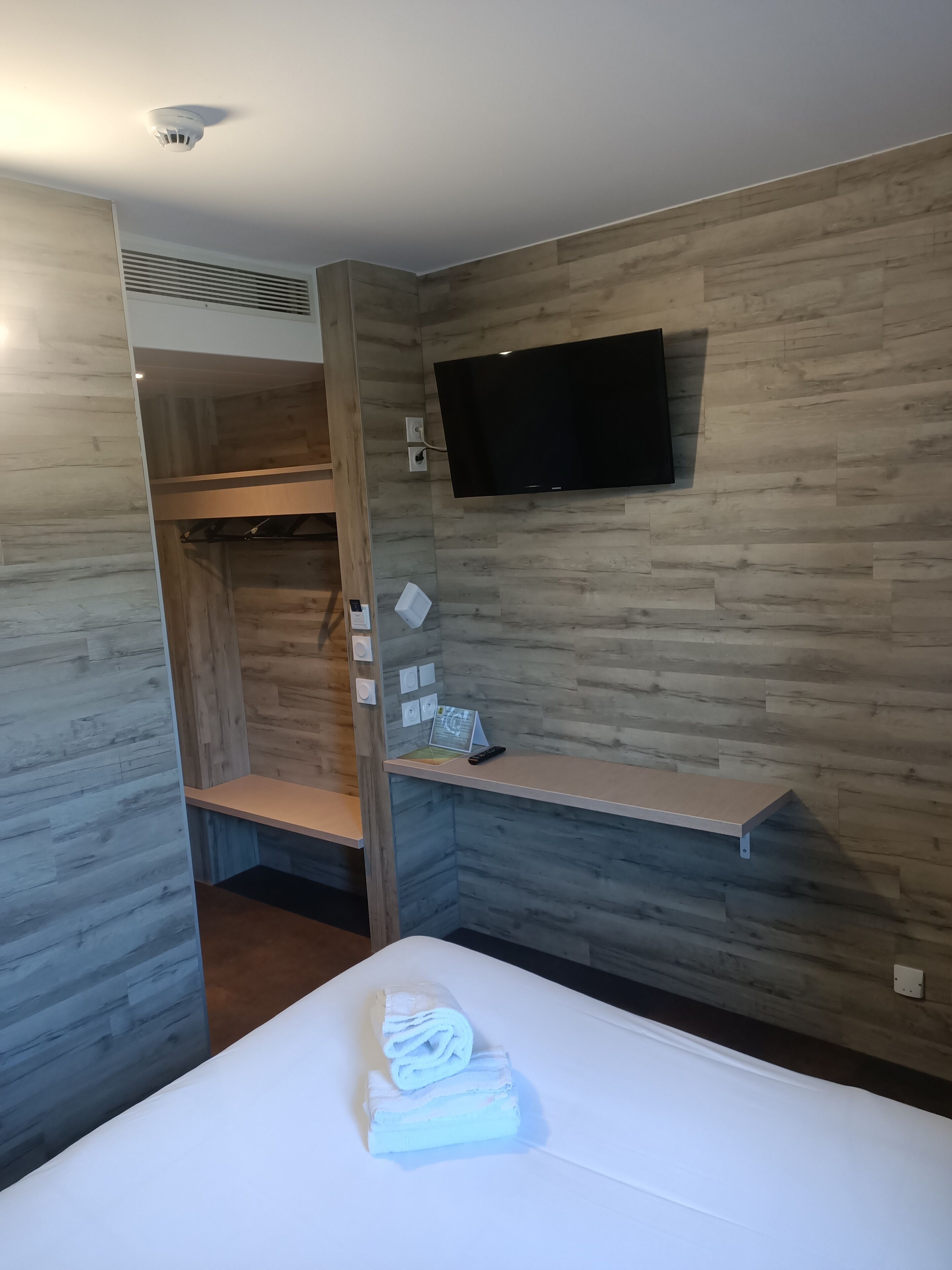 A hotel room with a bed, a TV mounted on the wall, a wooden wardrobe, and a small shelf with a remote control and a book.