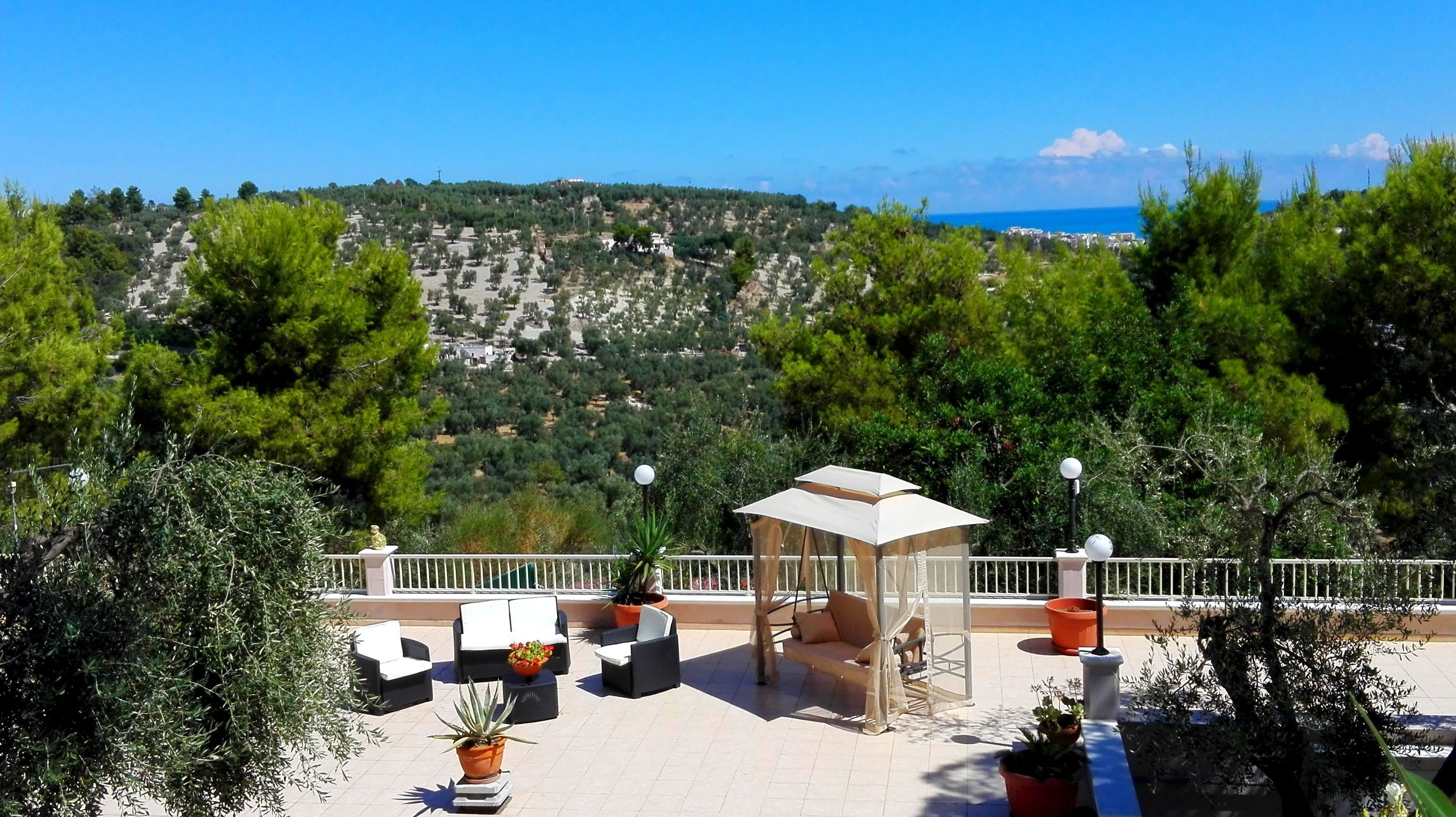 A terrace with outdoor seating, a small gazebo, and a view of a hilly landscape with trees and buildings in the distance.