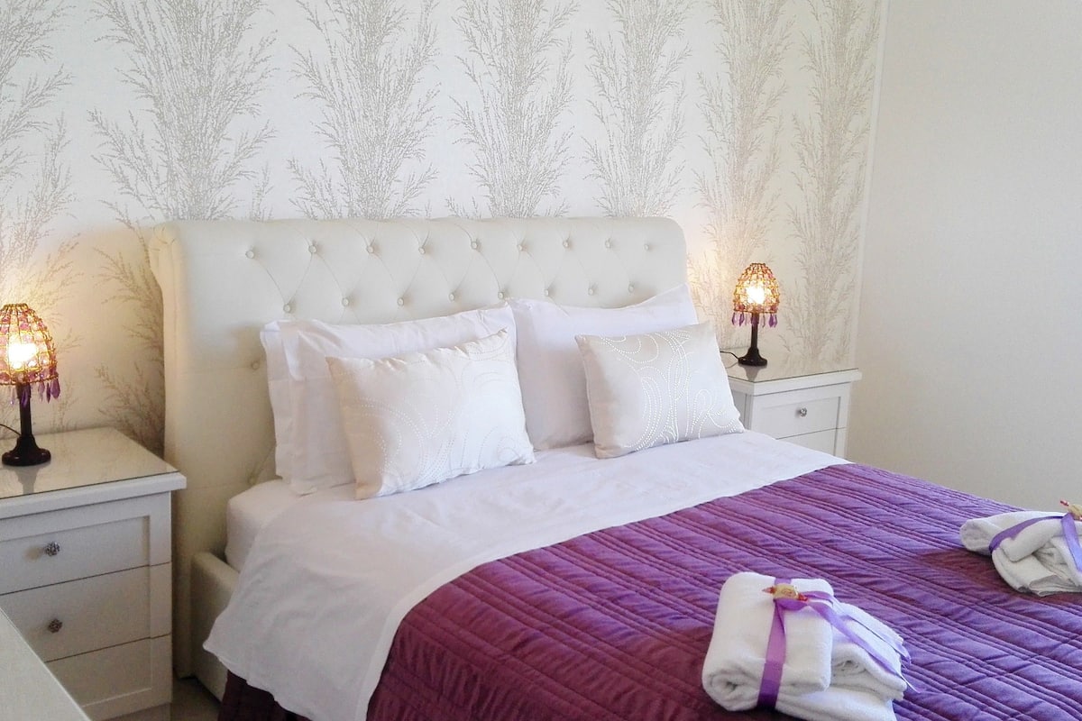Una cama bien hecha con colcha morada, almohadas blancas y cabecera blanca. Hay dos lámparas de noche sobre mesitas blancas.