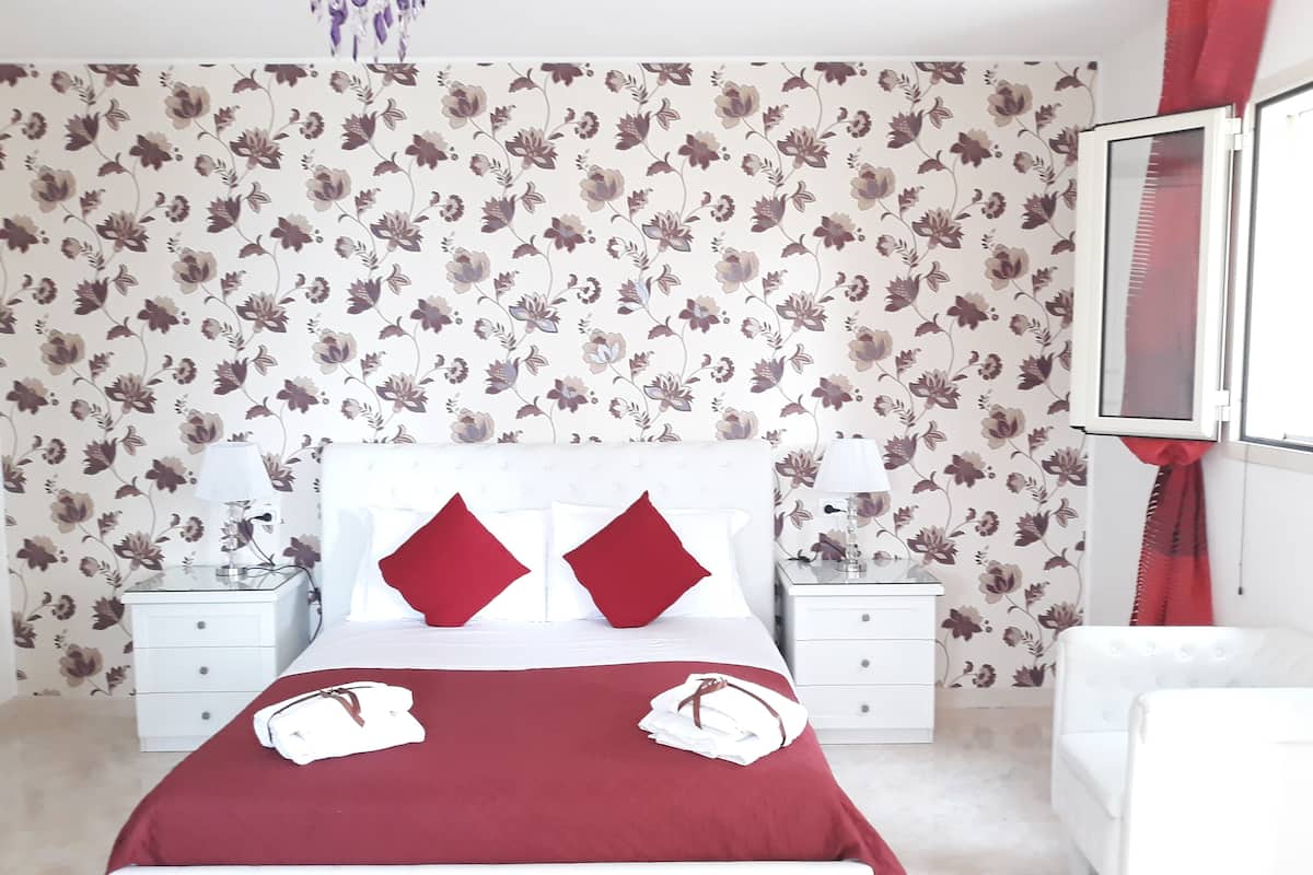 Un dormitorio con papel tapiz floral, una cama blanca con almohadas rojas, dos mesitas de noche con lámparas y una silla blanca.
