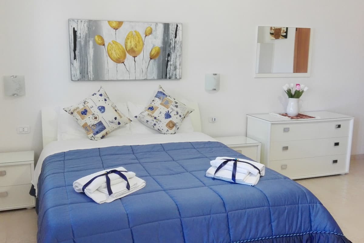 Una cama bien hecha con un edredón azul, dos almohadas con estampado floral, una mesita de noche con un florero y un cuadro de flores amarillas en la pared.