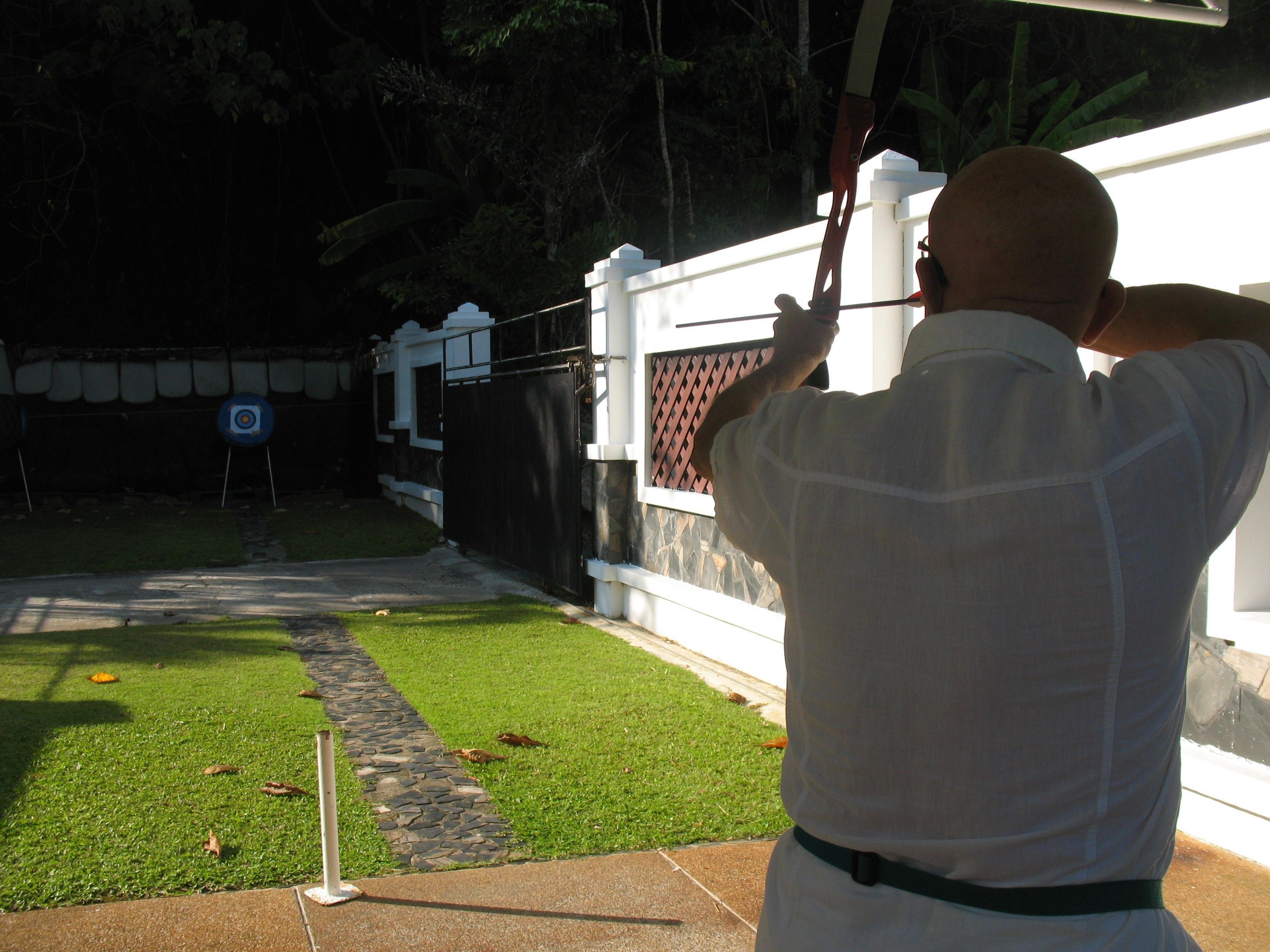 A man aiming a bow at a target in a backyard with a white fence and a stone pathway.