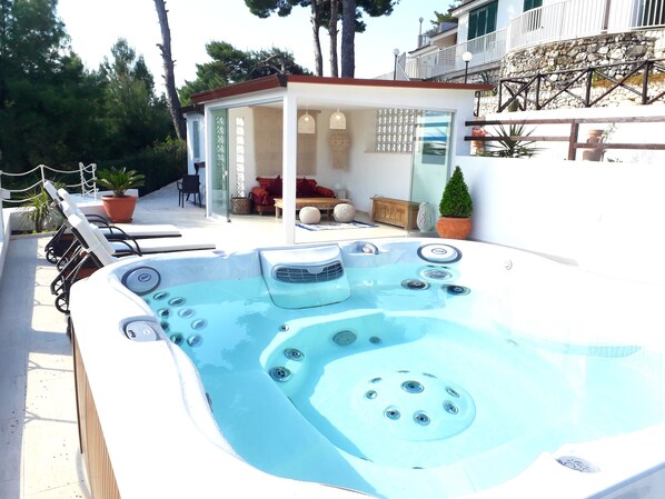 Un jacuzzi con múltiples chorros, un área de estar techada con cojines y una pared de piedra con plantas en macetas.