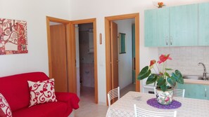 Un espacio habitable compacto con un sofá rojo, una mesa de comedor con un mantel individual morado y una zona de cocina con armarios color verde azulado.