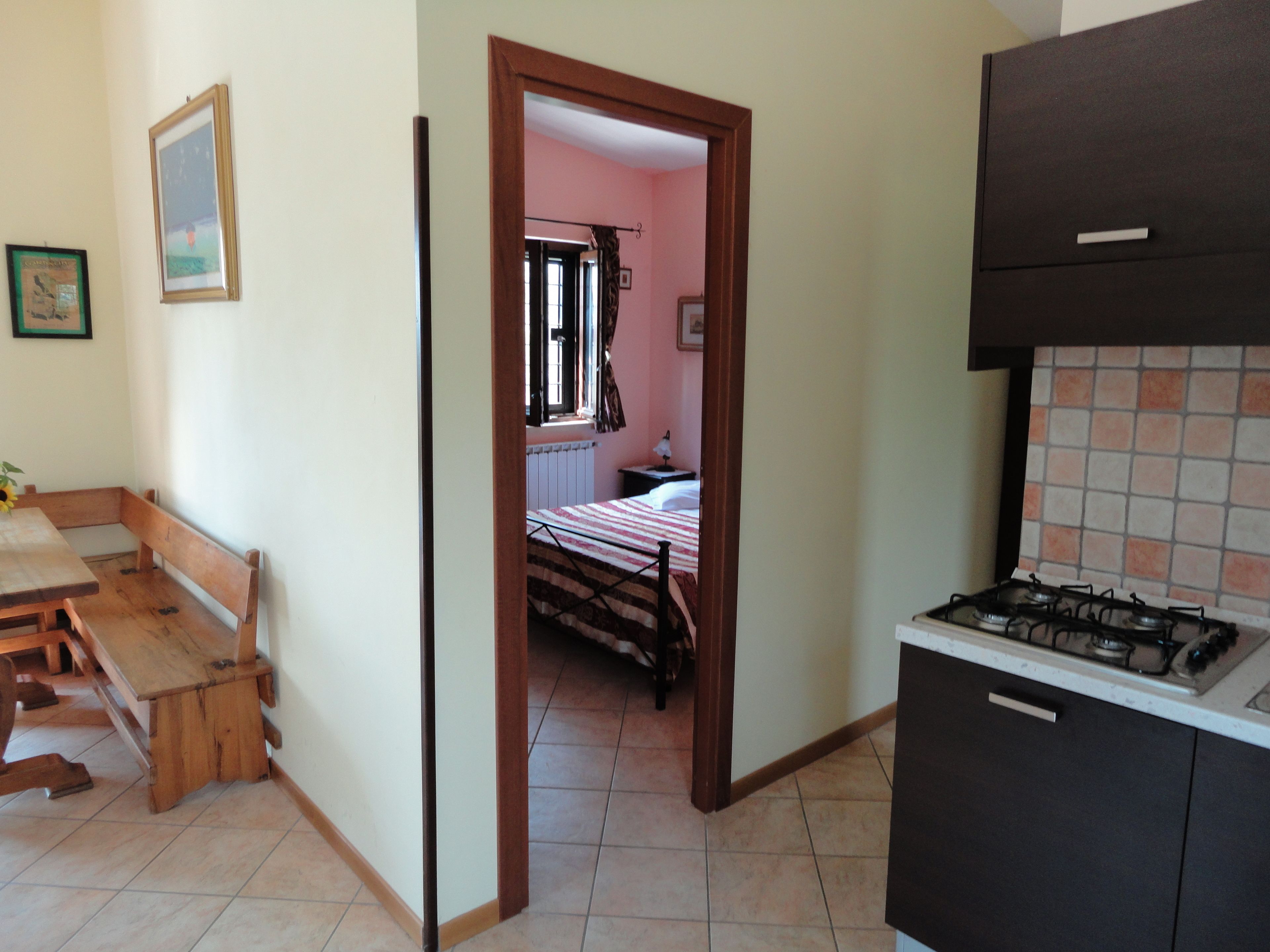 A kitchen with a gas stove, wooden bench, and a doorway leading to a bedroom with a bed and window.