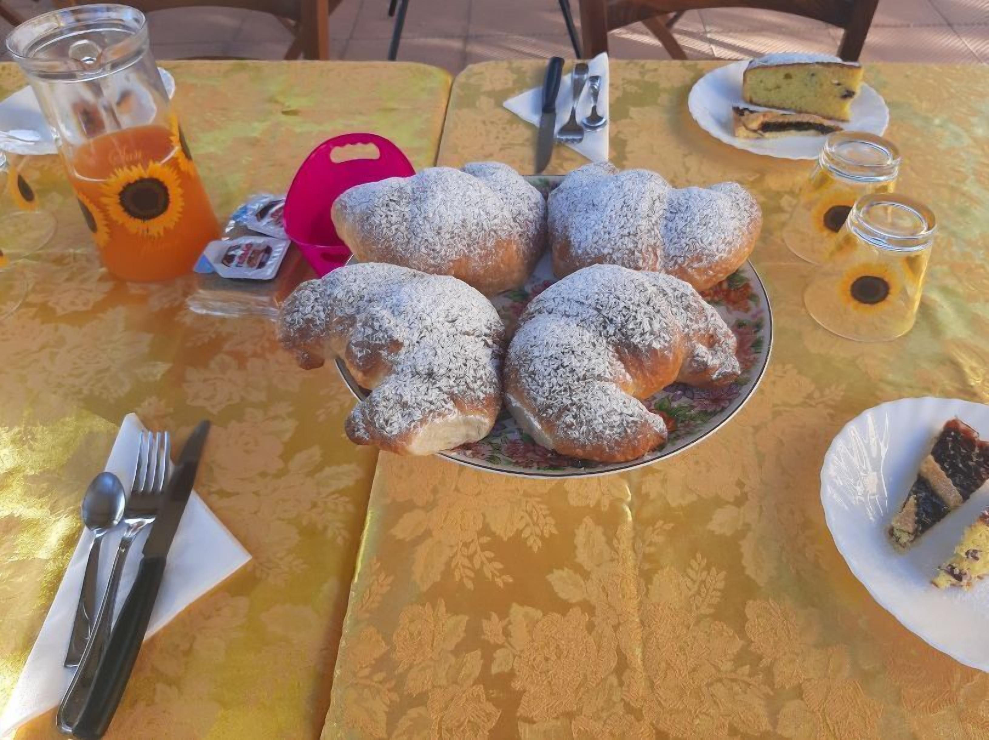 A table with a plate of pastries dusted with powdered sugar, a glass of orange juice, and a slice of cake on a floral tablecloth.