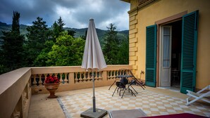 A balcony with a checkered floor, a table with chairs, a red lounge chair, and a potted plant with red flowers. - Marta Guest House (Lucca)