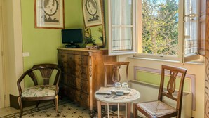 A room with a green wall, a wooden cabinet, two chairs, a small table with a vase, and a window with a view of trees. - Marta Guest House (Lucca)