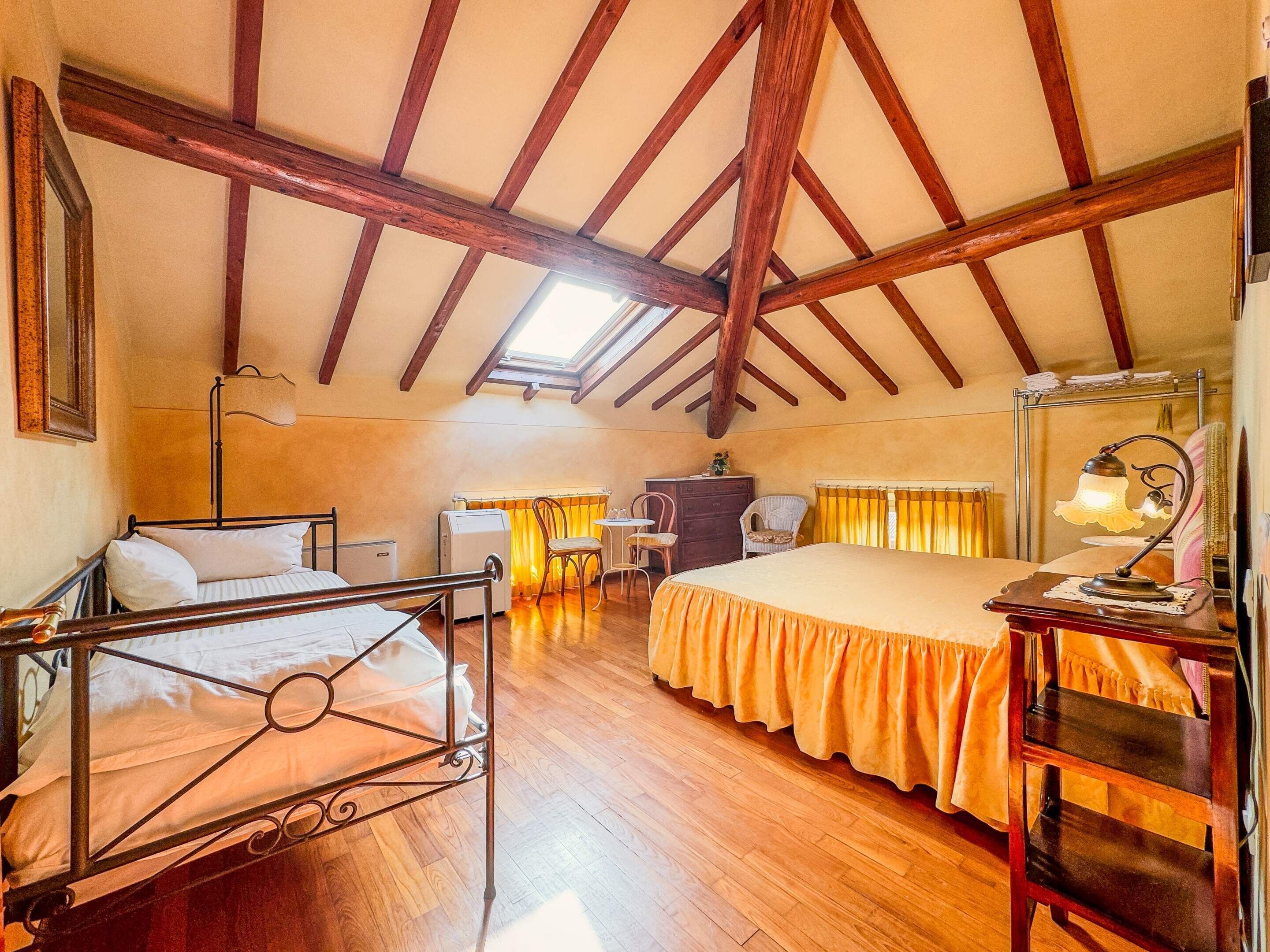 A room with a sloped ceiling, wooden beams, a bed with a yellow bedspread, a desk with a lamp, and a chair.