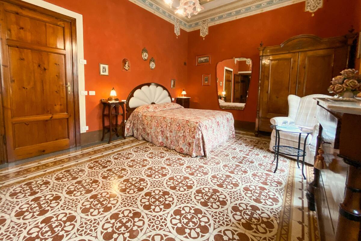 A bedroom with a large wooden door, a bed with a floral bedspread, a chair, a small table, and a mirror.