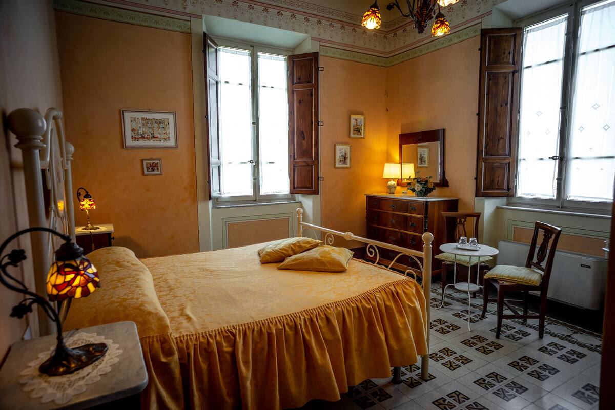A bedroom with a bed, a bedside table with a lamp, a dresser, a chair, and a window with shutters.