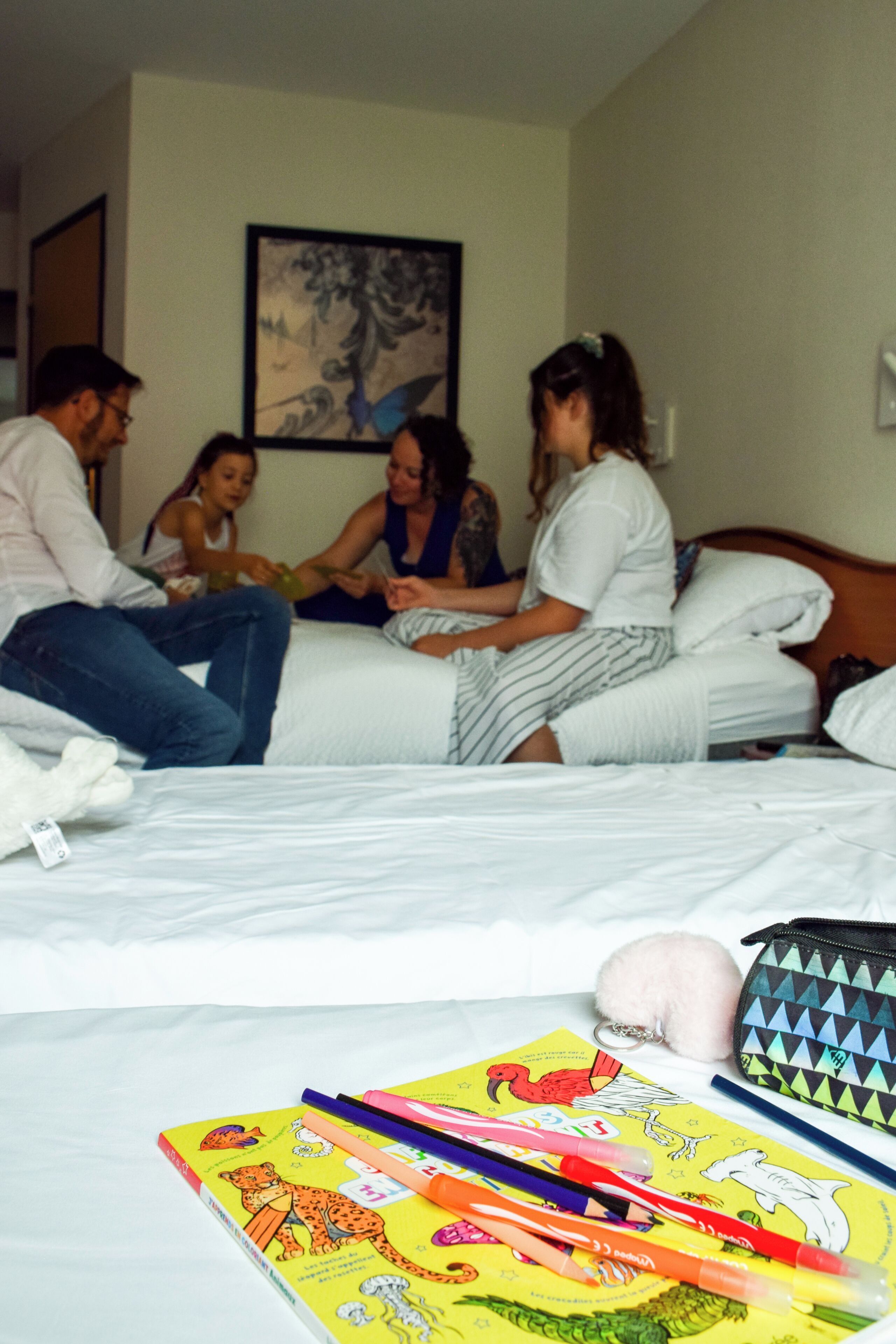 A room with a bed, a painting on the wall, and a group of people sitting on the bed.