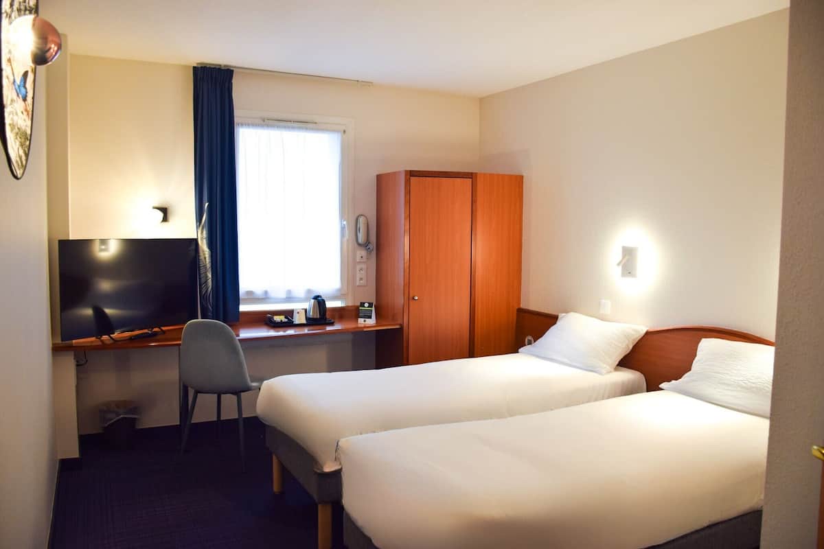 Une chambre d’hôtel avec deux lits, un bureau avec un ordinateur, une chaise, une armoire et une fenêtre avec des rideaux.