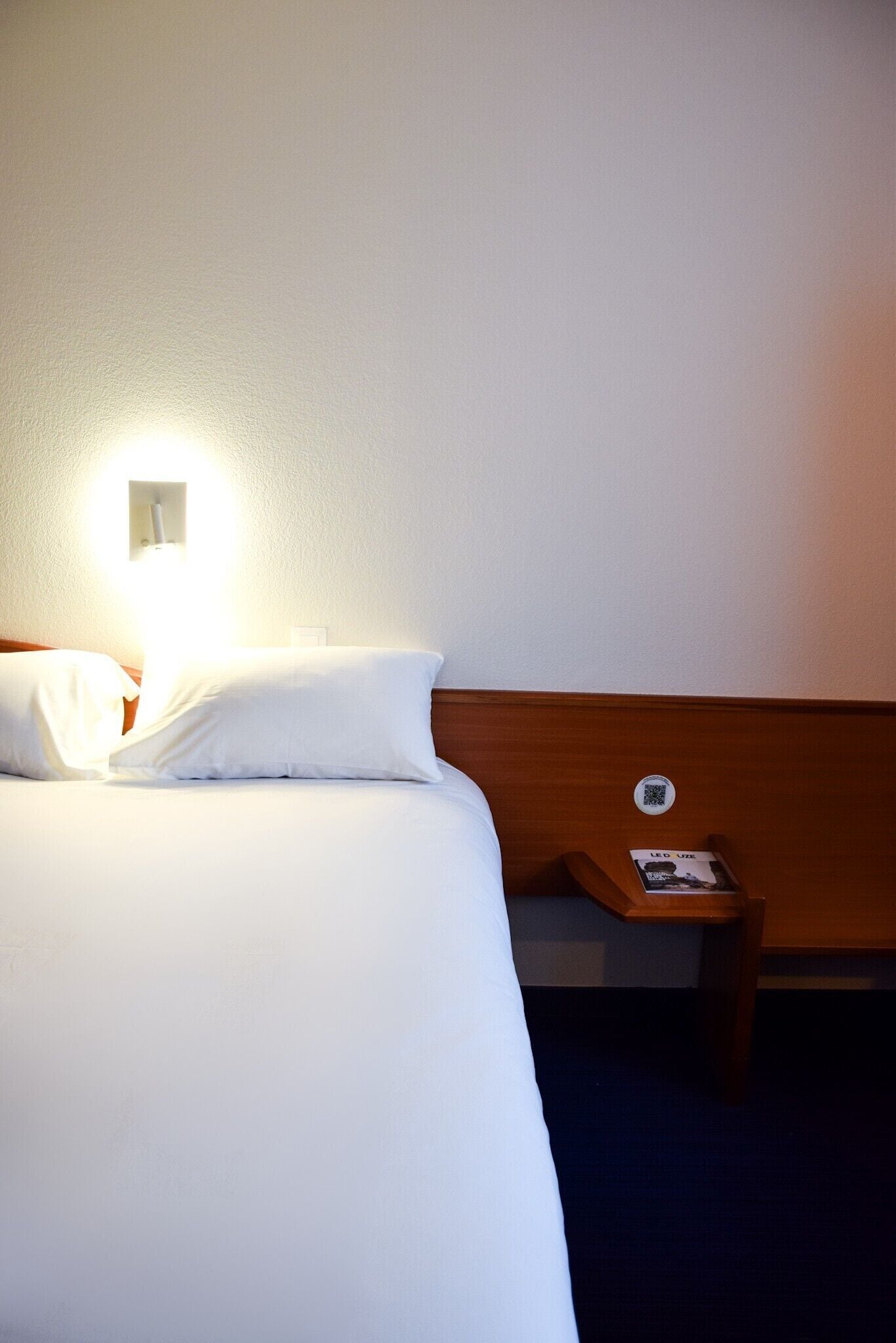 A hotel room with a neatly made bed, a bedside table with a lamp, and a wall-mounted control panel.