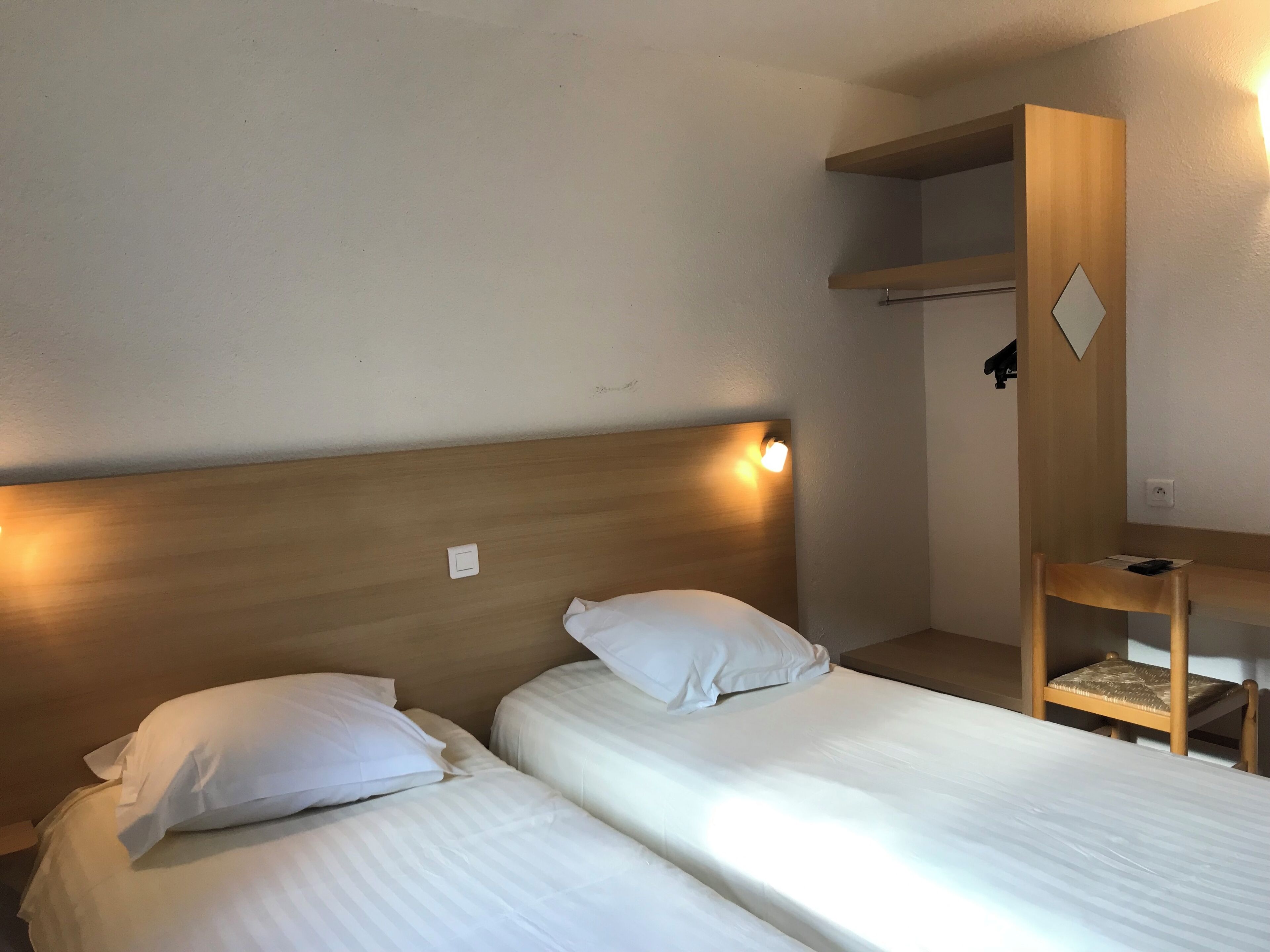 Ein Hotelzimmer mit zwei Einzelbetten, einem Holzkopfteil, einem Nachttisch, einem Schreibtisch und einem Regal.