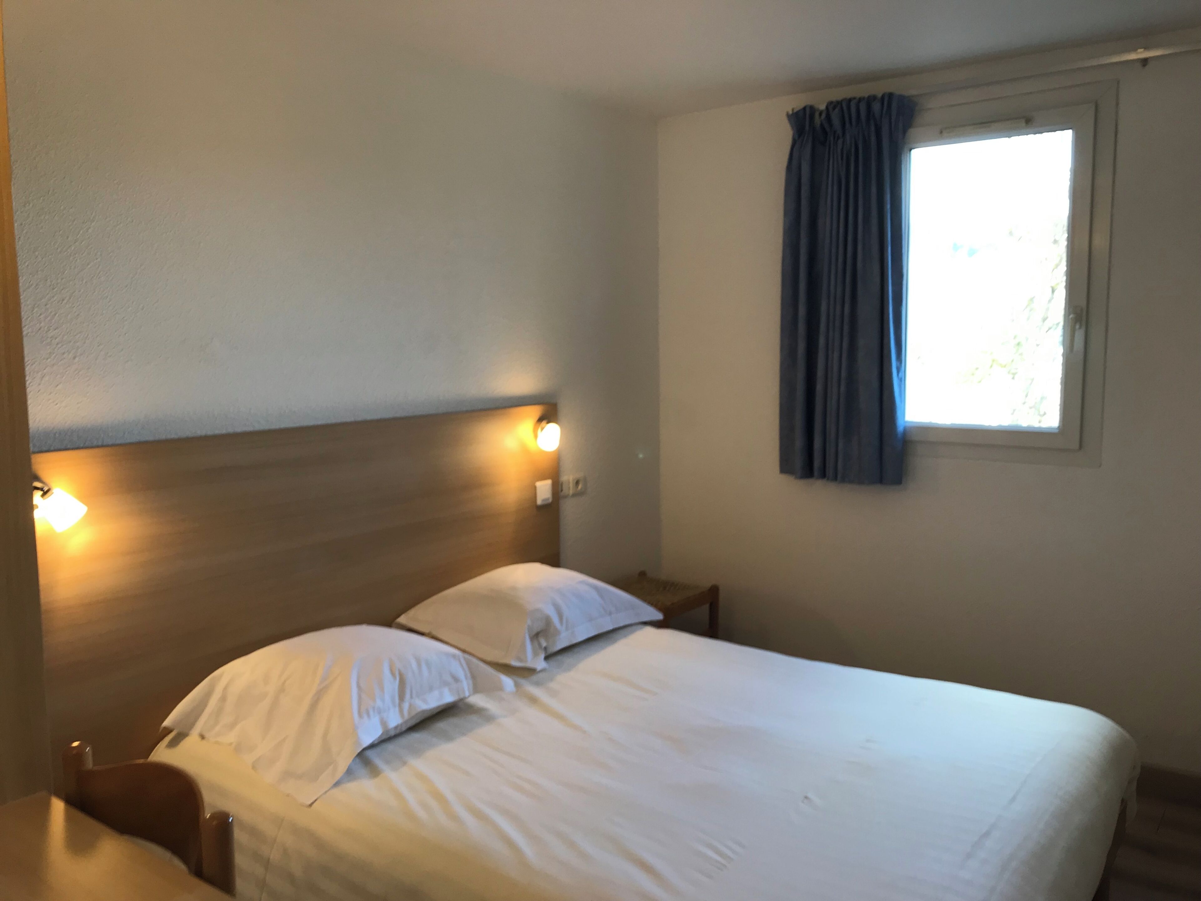 A single bed with white linens, a wooden headboard, two bedside lamps, and a window with a curtain.