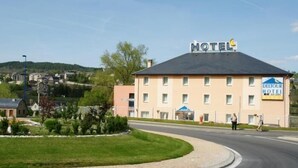 Um hotel com uma placa indicando ser um DEUTOUR HOTEL, apresentando um edifício moderno com telhado plano e um amplo estacionamento.