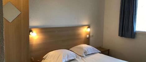 A single bed with a wooden headboard, a bedside table, and a window with curtains.