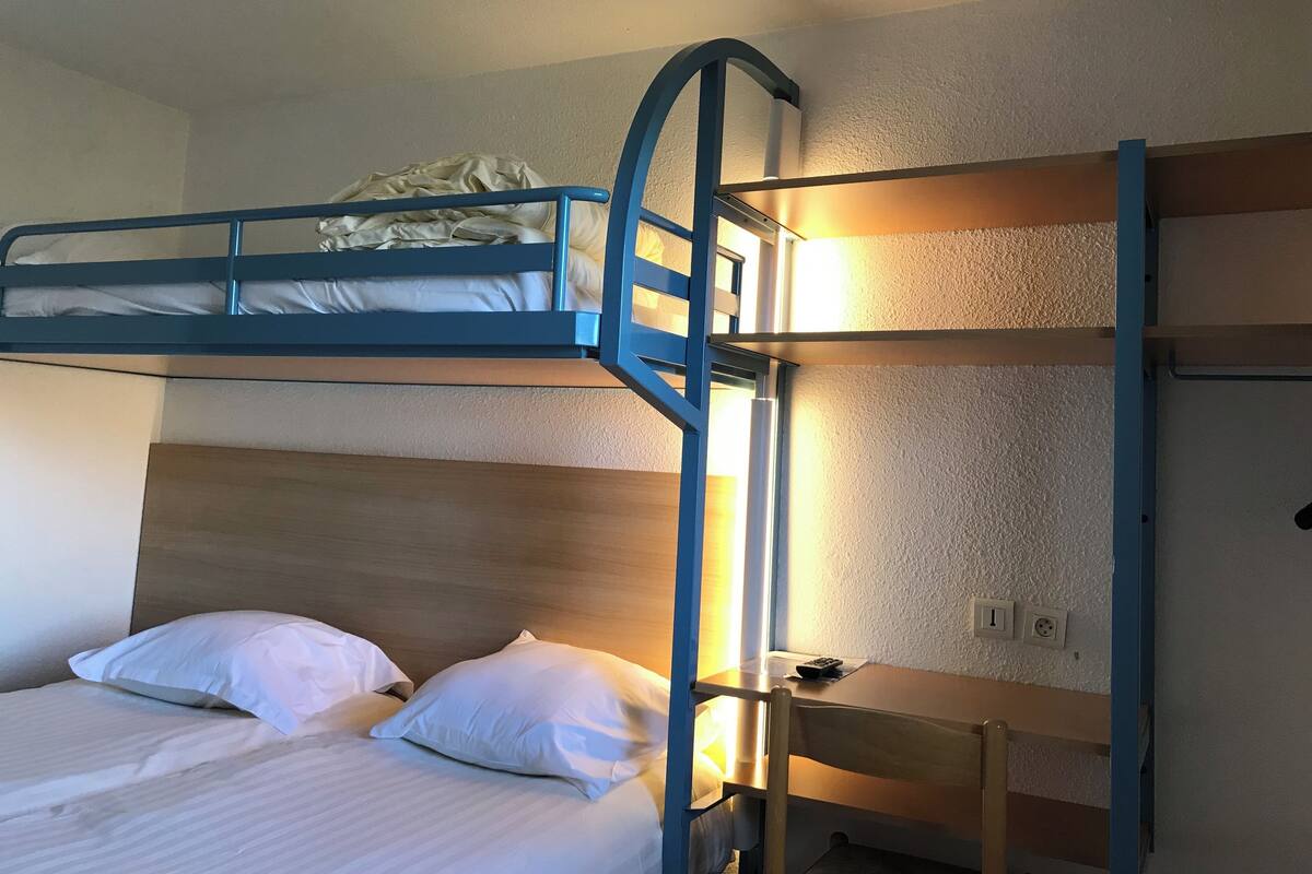 A bunk bed with a ladder, a desk with a chair, and a bedside table with a remote control.