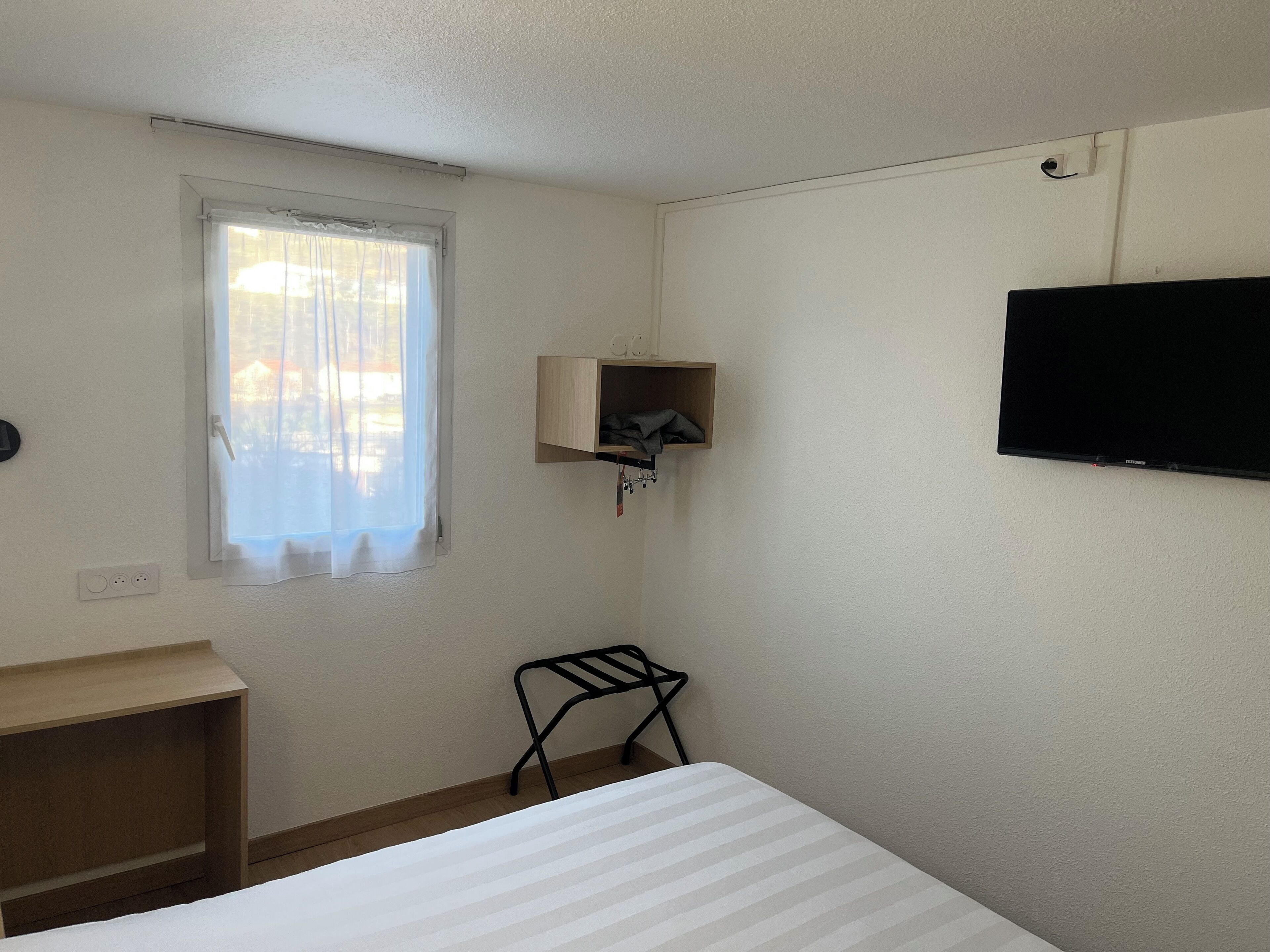 A small, clean room with a bed, a TV, a window with curtains, a small shelf, and a chair.