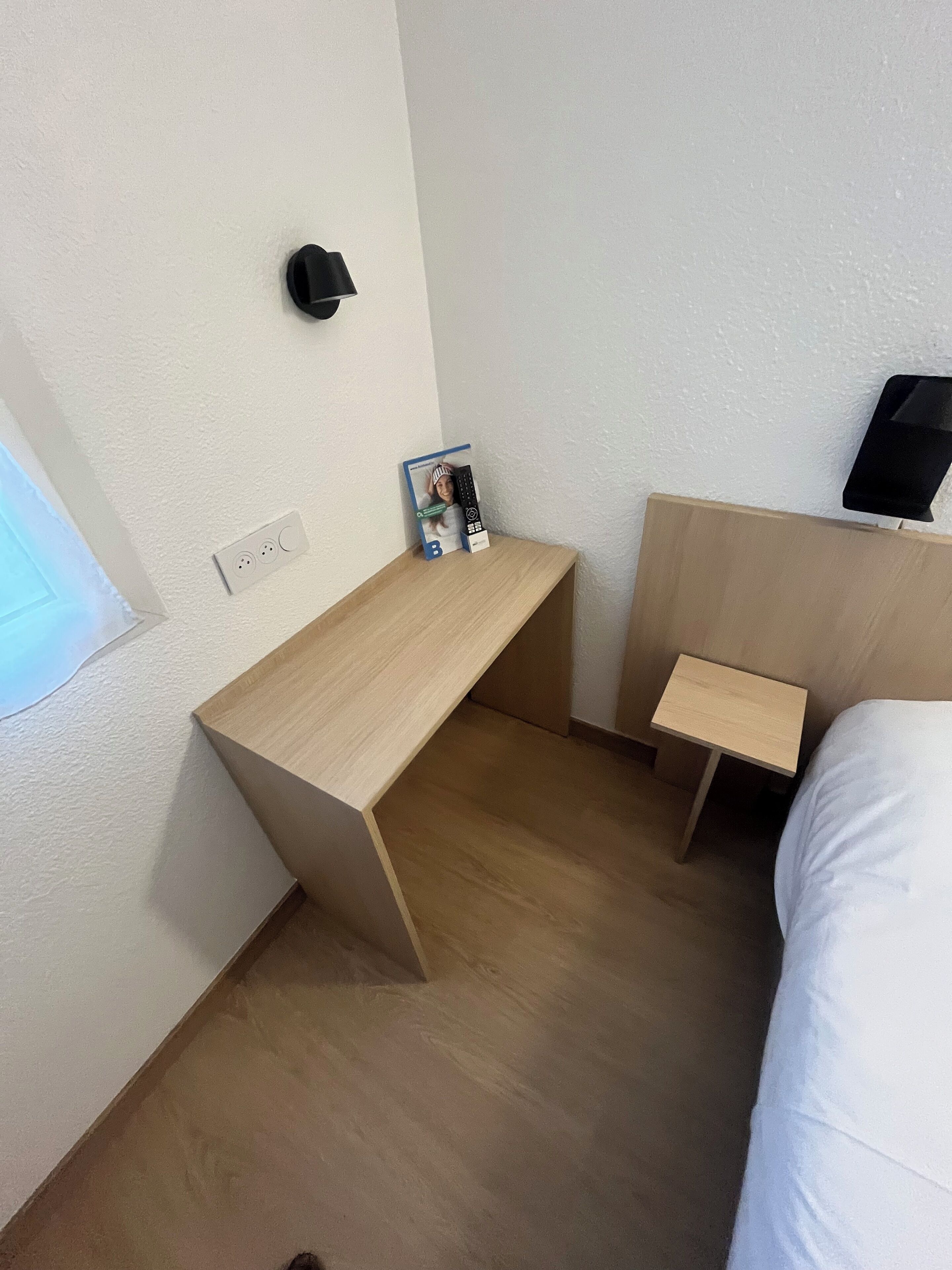 Een kleine hotelkamer met een houten bureau, nachtkastje, bed en een aan de muur bevestigde televisie.