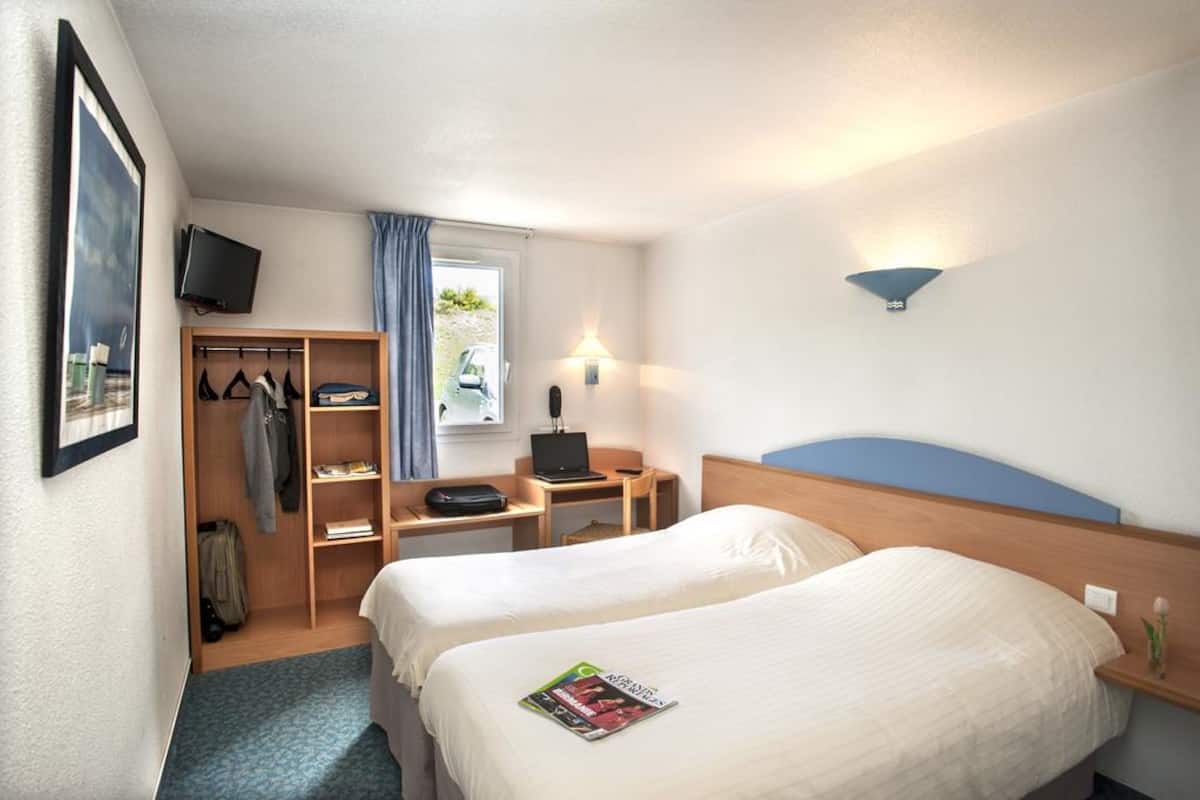 Ein Hotelzimmer mit zwei Betten, einem Schreibtisch mit einem Laptop, einem an der Wand befestigten Fernseher und einem Kleiderschrank mit Kleidung.