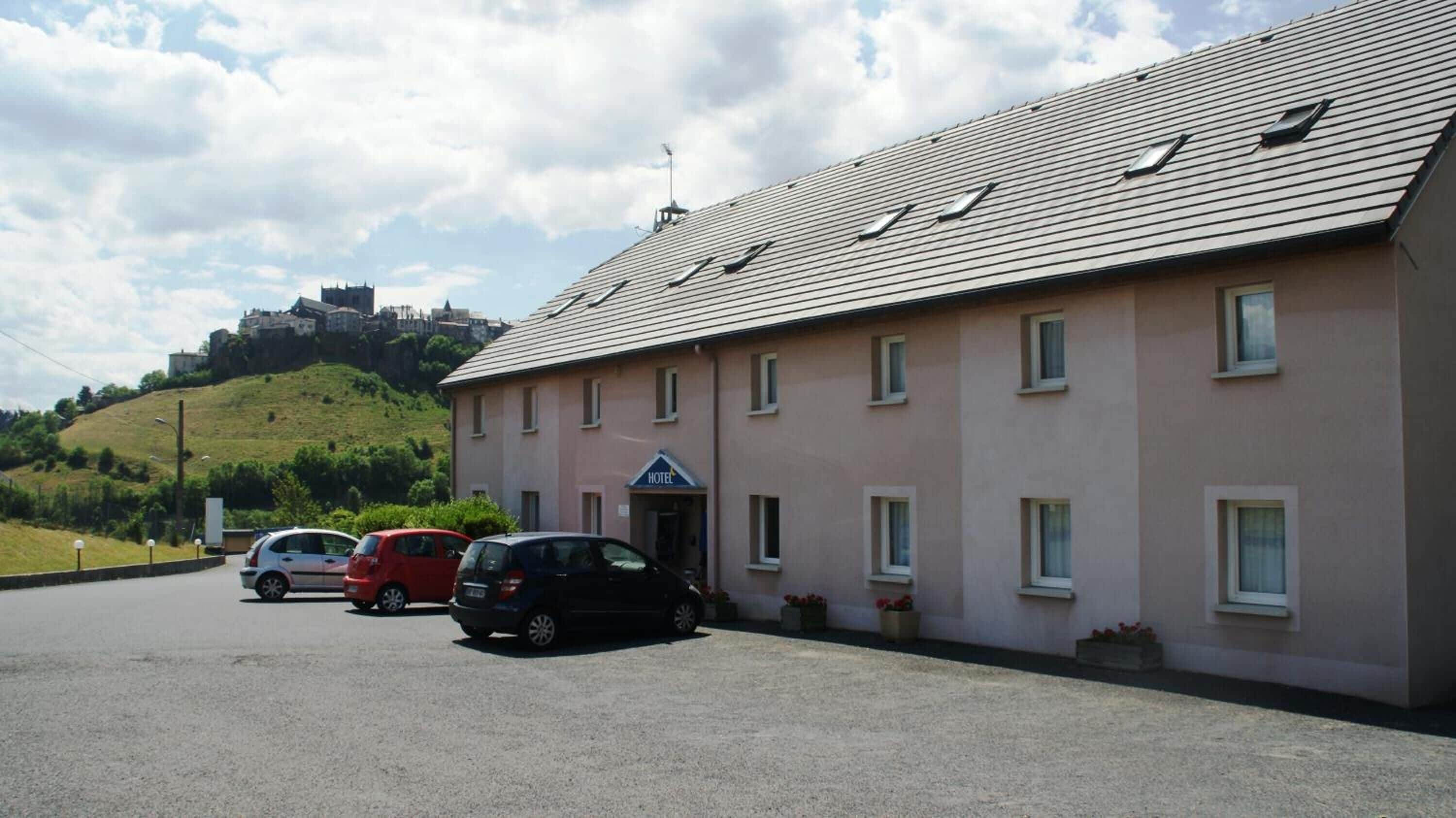 A hotel with a flat roof, multiple windows, and a sign indicating it is a hotel. There are cars parked in front, and a hill with a castle-like structure in the background.