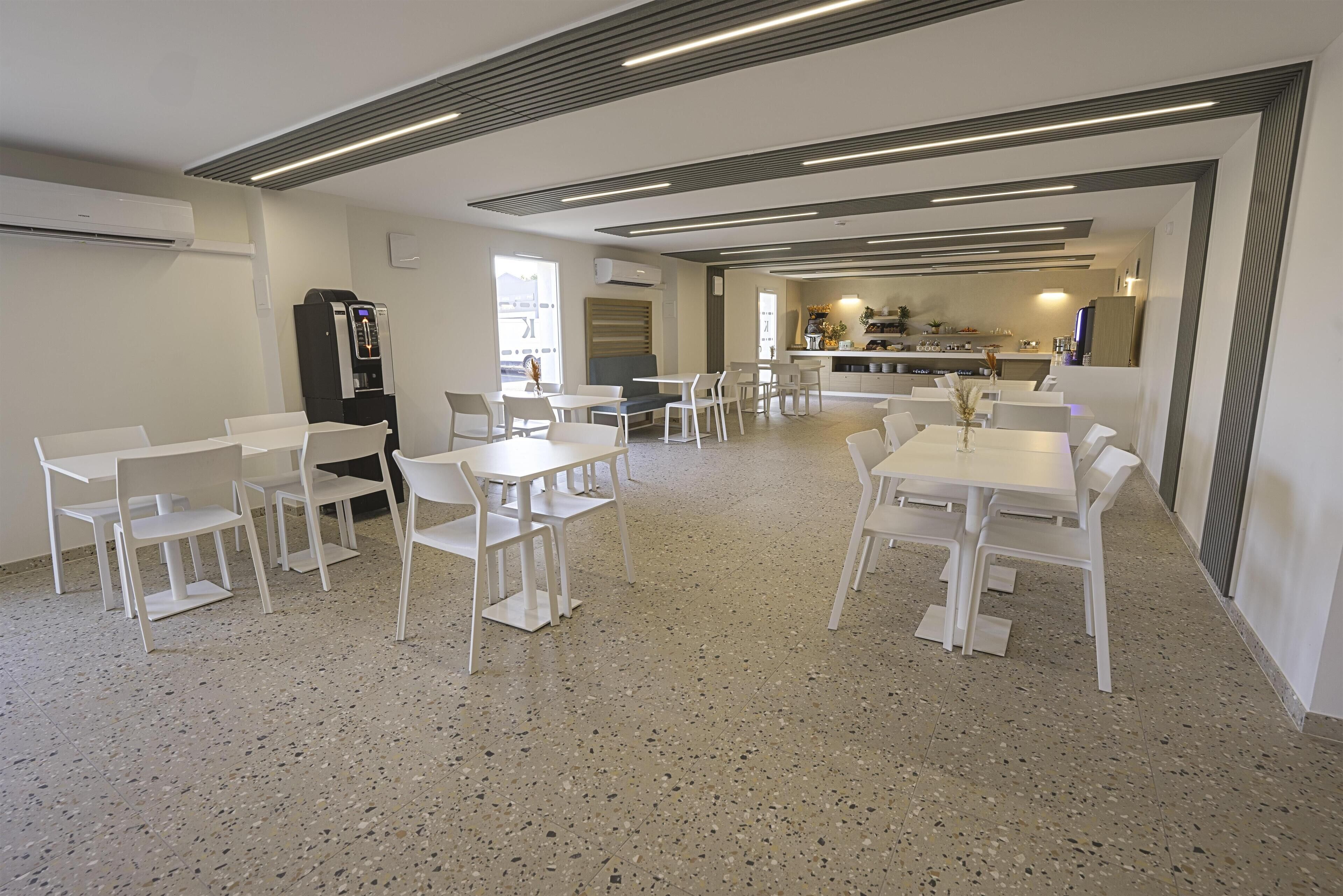 Un grand espace dédié à la salle à manger, avec des tables et des chaises blanches, un coin cuisine à l’arrière-plan et un sol à motifs.