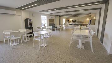 A spacious dining area with white tables and chairs, a kitchenette in the background, and a speckled floor.