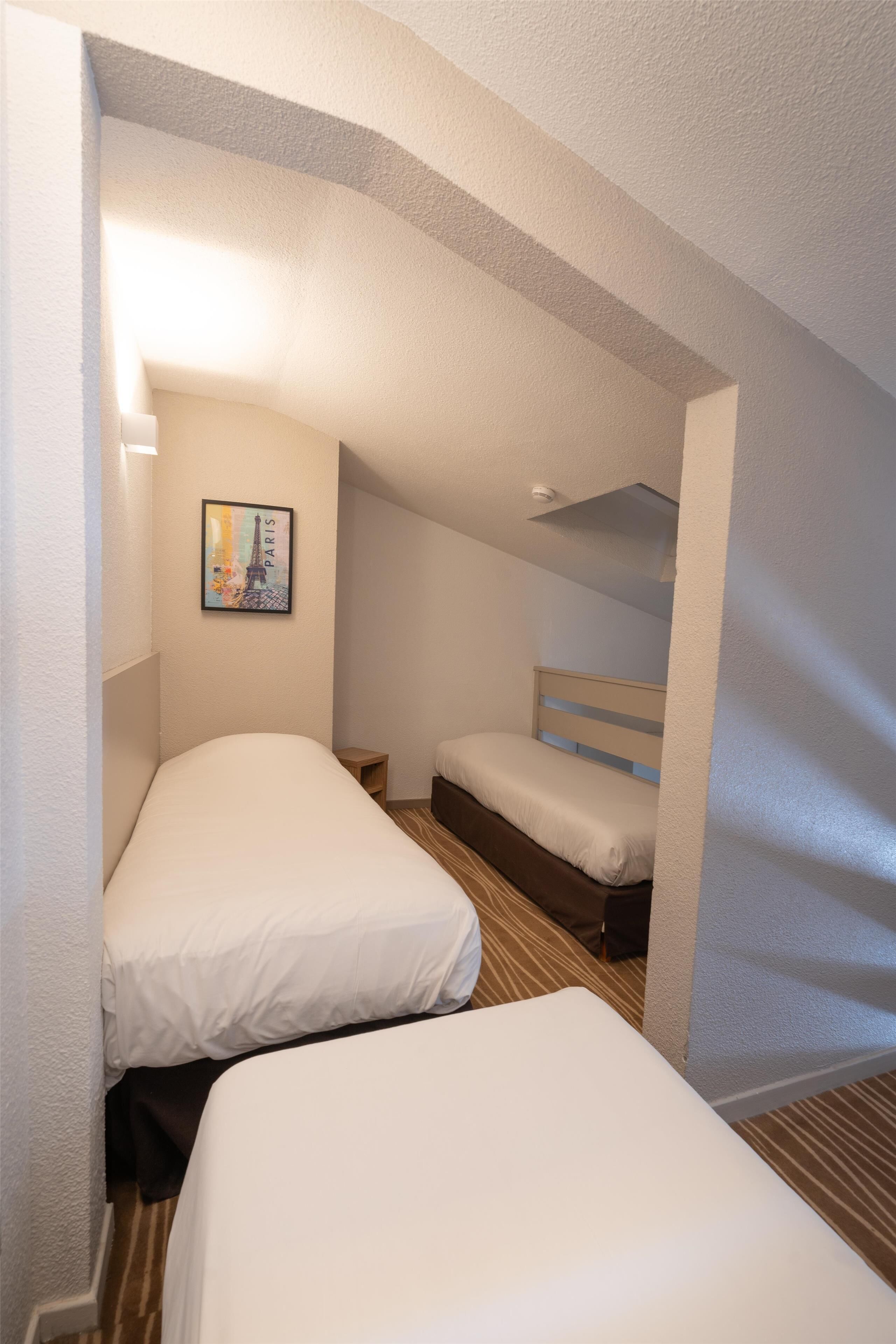 A compact hotel room with two single beds, a small bedside table, a wall-mounted picture of the Eiffel Tower, and a sloped ceiling.