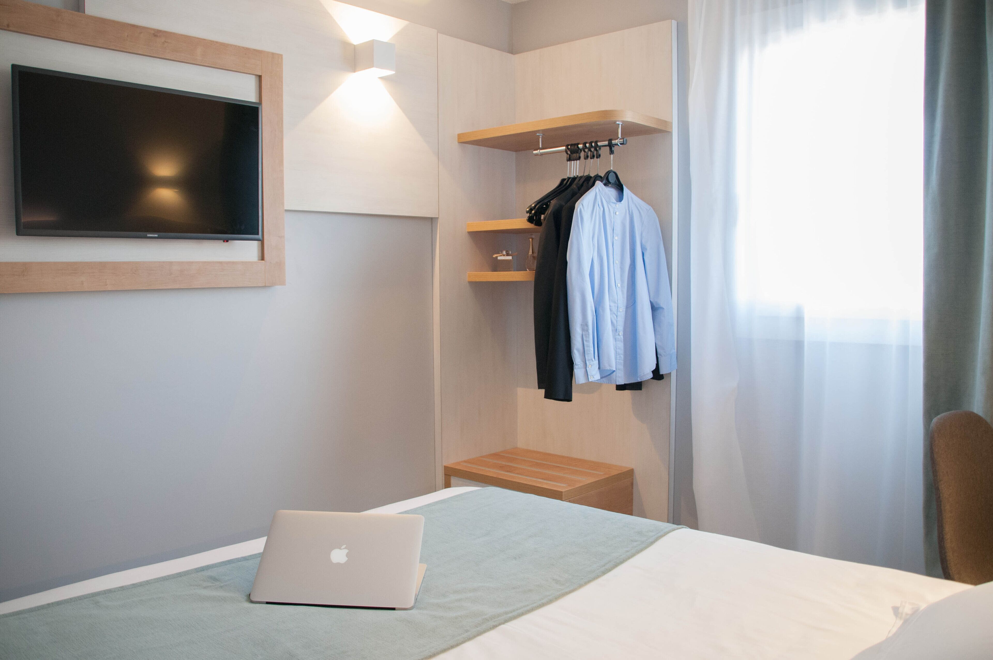 Ein Hotelzimmer mit Bett, Laptop, Wandfernseher, einem Holzregal mit Kleidung und einem Fenster mit Vorhängen.