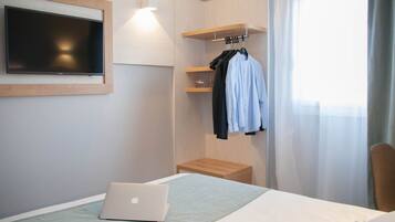 A hotel room with a bed, a laptop, a TV mounted on the wall, a wooden shelf with clothes, and a window with curtains.