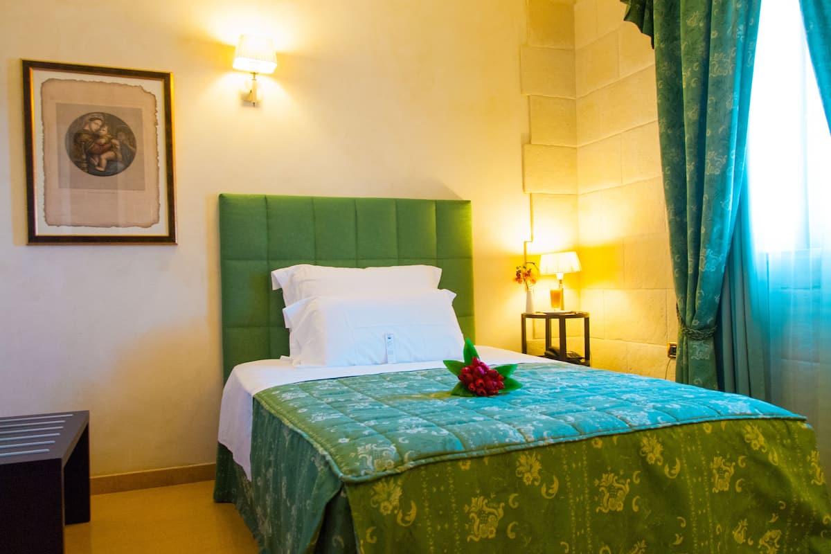 A neatly made bed with a green headboard, white bedding, and a decorative flower arrangement. There are two wall-mounted lamps and a framed picture on the wall.