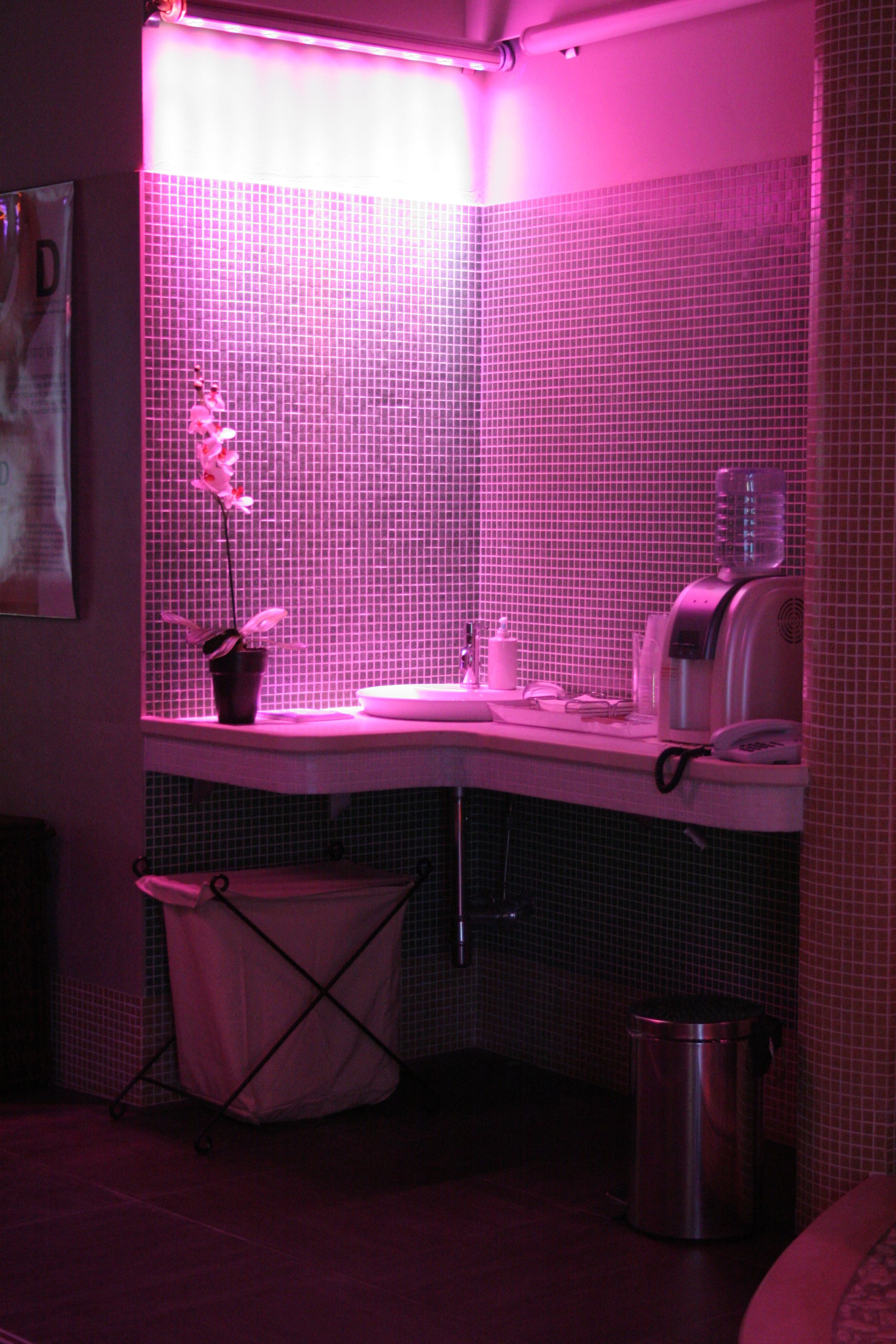 Un baño con un lavabo iluminado en rosa, un cubo de basura y un dispensador de agua.