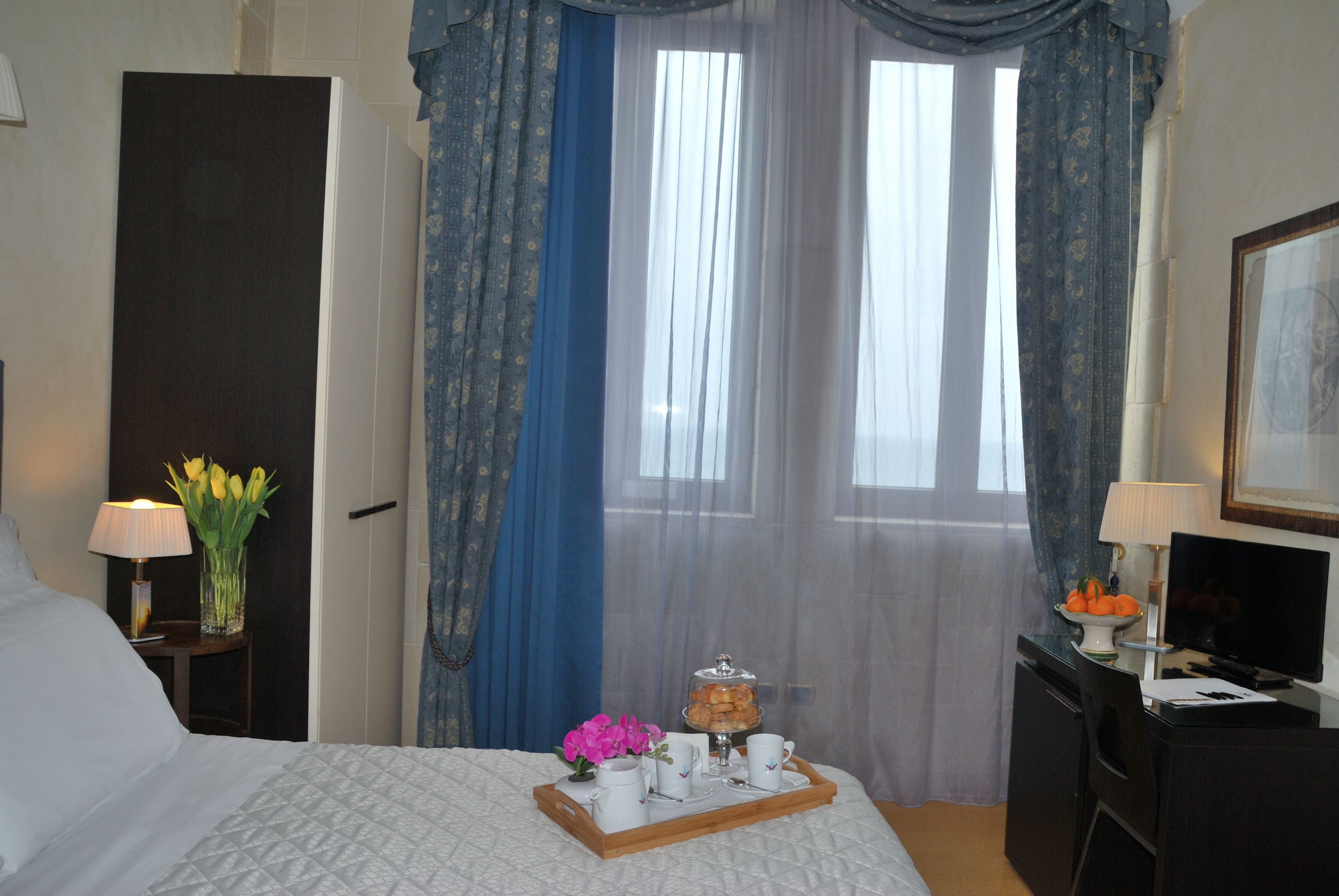 A hotel room with a bed, a desk with a computer, a nightstand with a lamp, and a window with curtains.