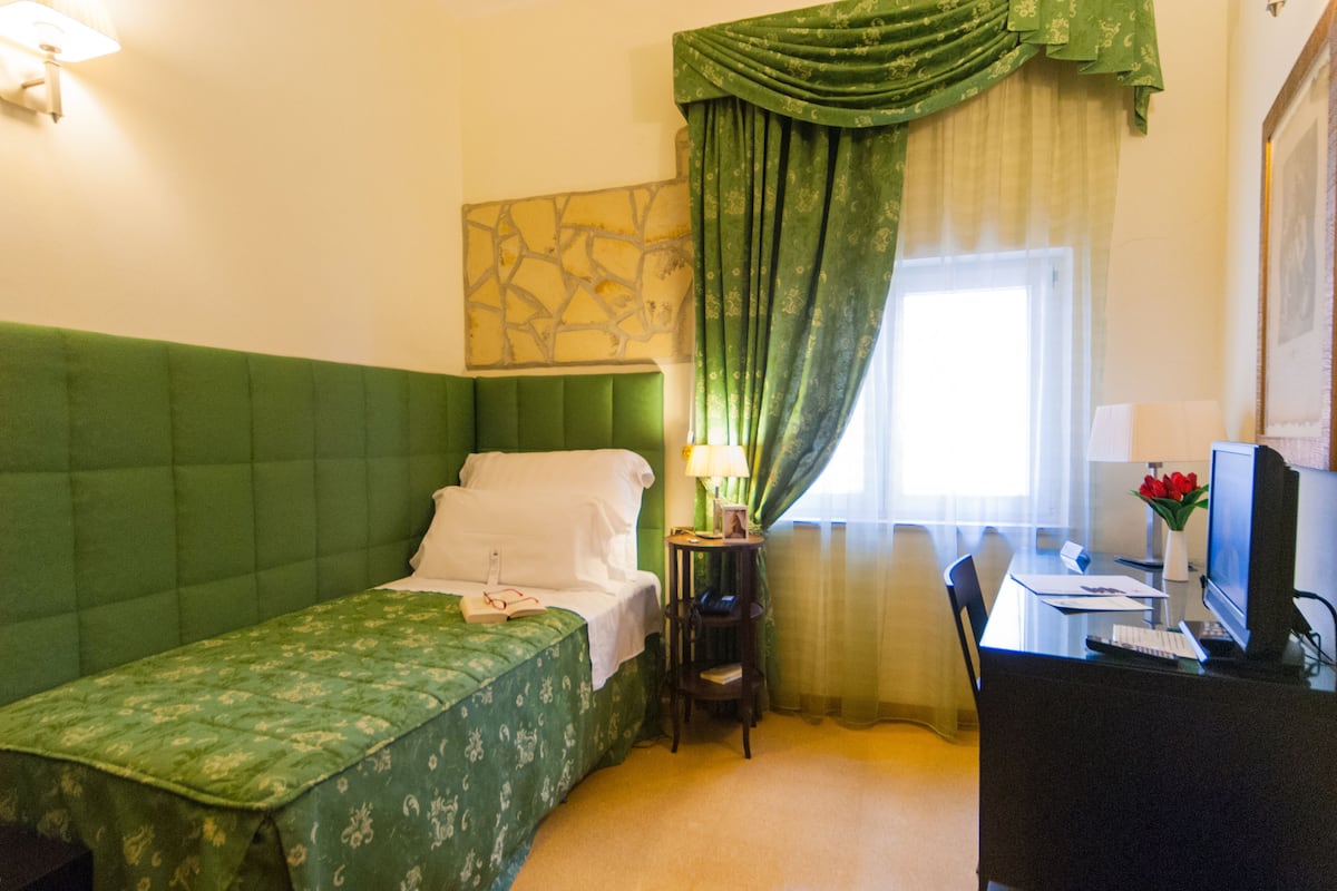 A hotel room with a green bed, a desk with a computer, a chair, and a window with curtains.