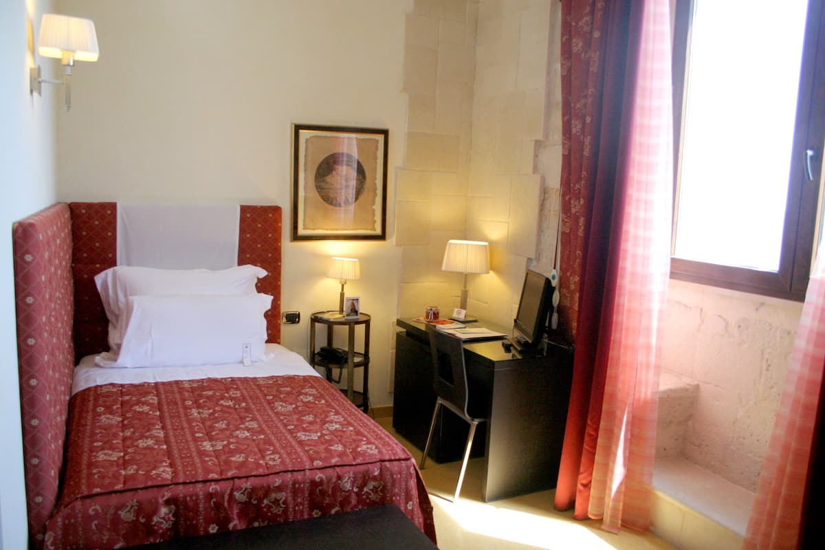A hotel room with a bed, desk, and computer, a framed picture on the wall, and a window with curtains.