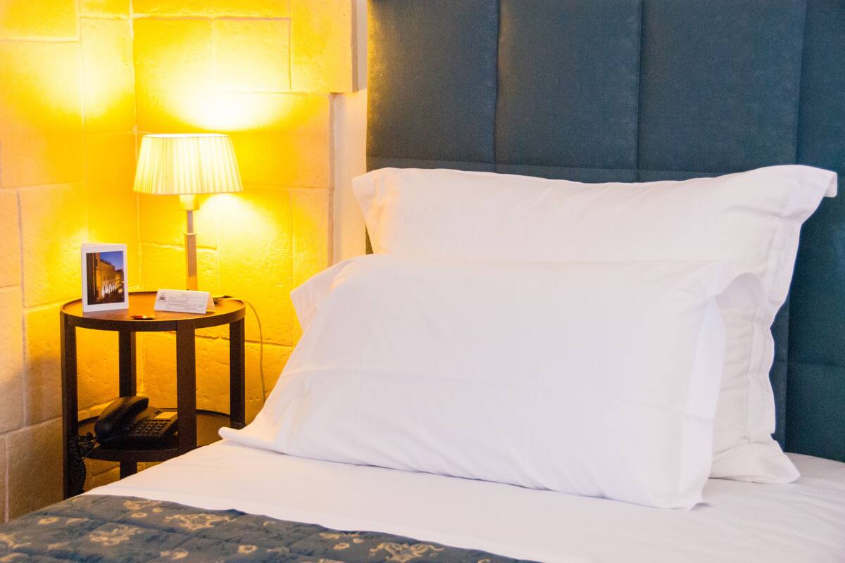 A neatly made bed with white linens, a bedside lamp, a small table with a phone and a picture frame, and a wall with a textured pattern.