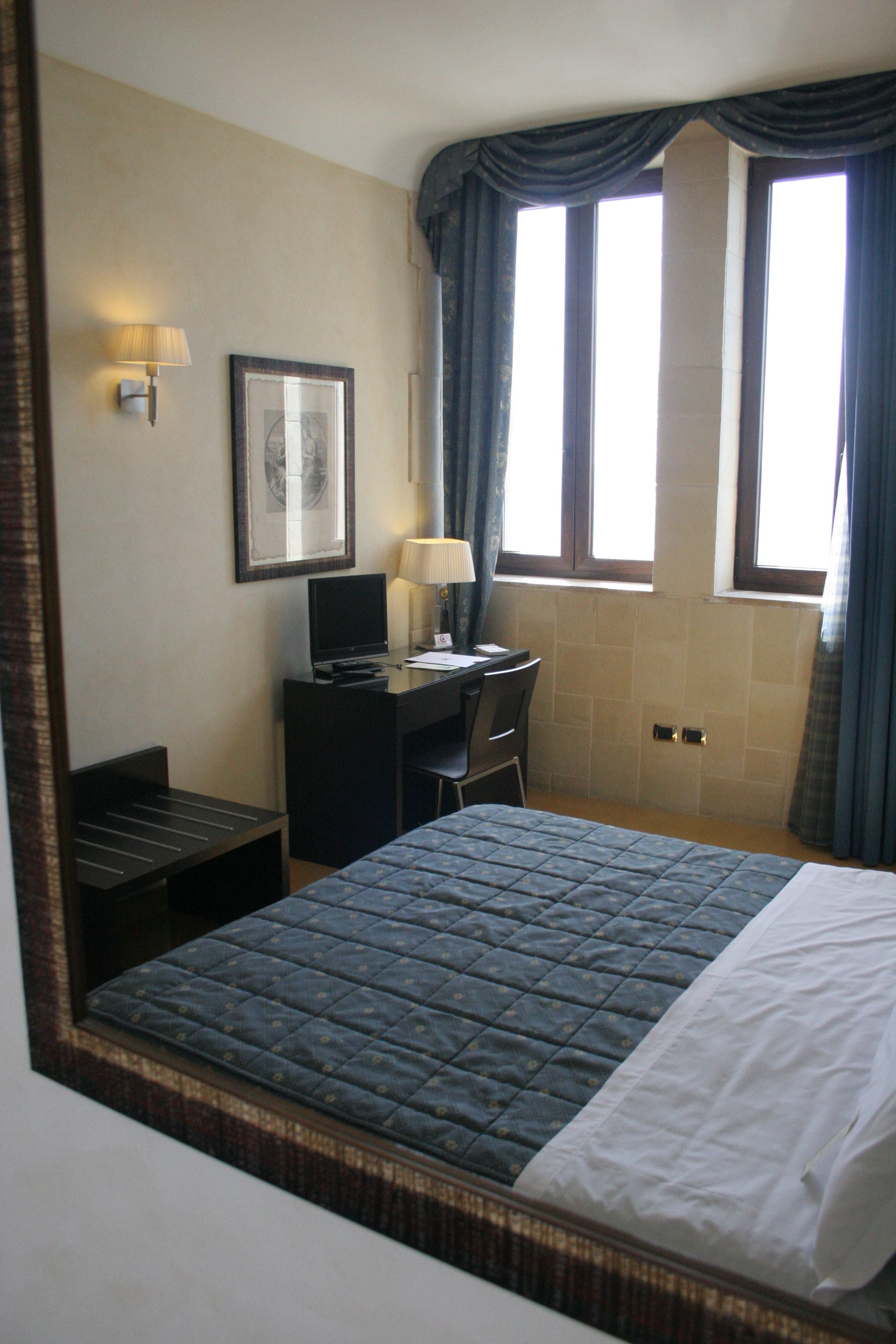Ein Hotelzimmer mit Bett, Schreibtisch mit Computer, Stuhl, Lampe und einem gerahmten Bild an der Wand.