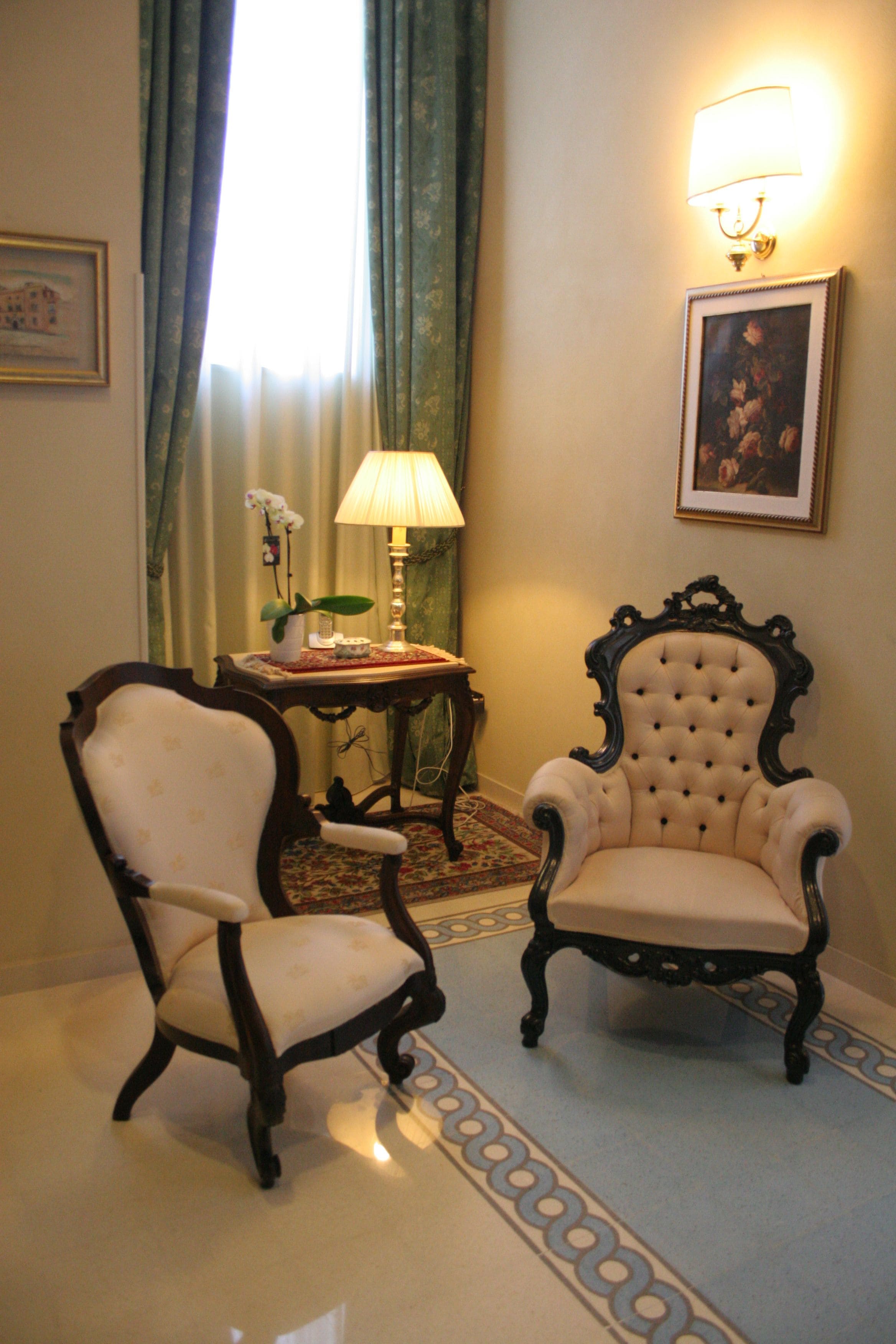 A room with two upholstered chairs, a table with a lamp, a framed picture on the wall, and a window with curtains.