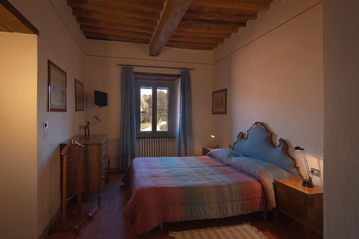 A bedroom with a wooden ceiling, a bed with a blue headboard, a nightstand with a lamp, and a window with curtains.