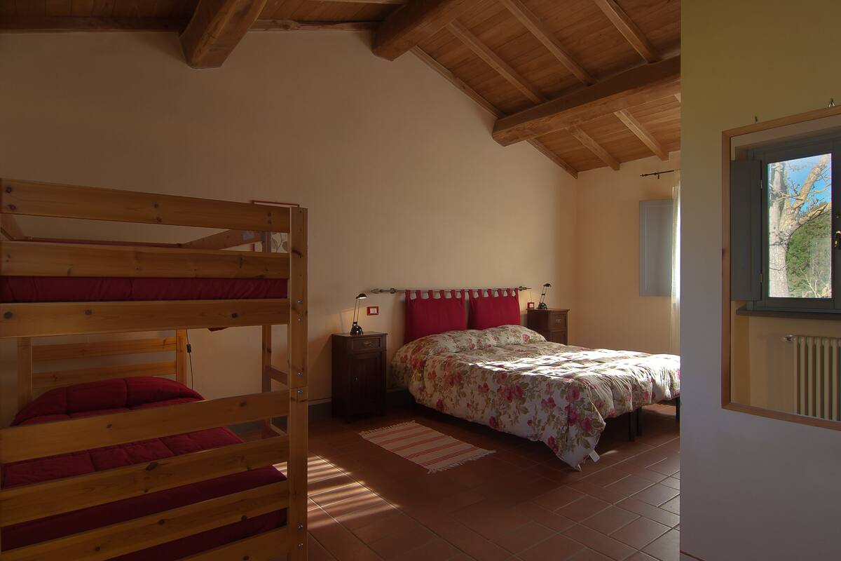 A bedroom with a bunk bed, a single bed with floral bedding, a nightstand, and a window with a view of trees.