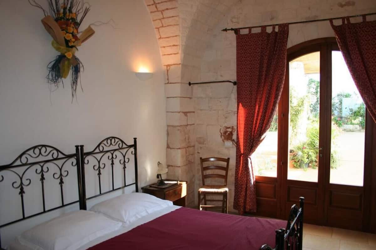 A bedroom with a bed, a chair, a nightstand, a wall-mounted light, and a window with curtains.