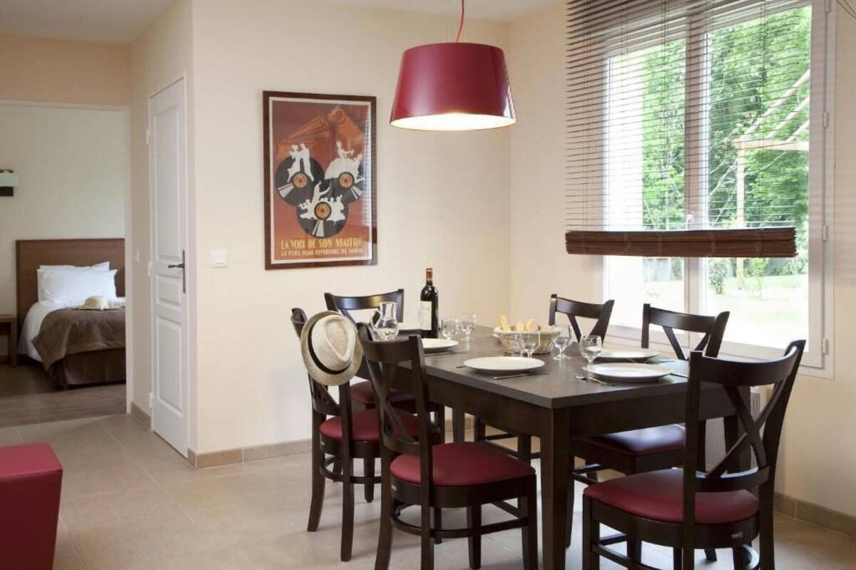 A dining area with a table set for four, surrounded by dark wooden chairs with red cushions. A red pendant light hangs above the table, and a poster is visible on the wall.